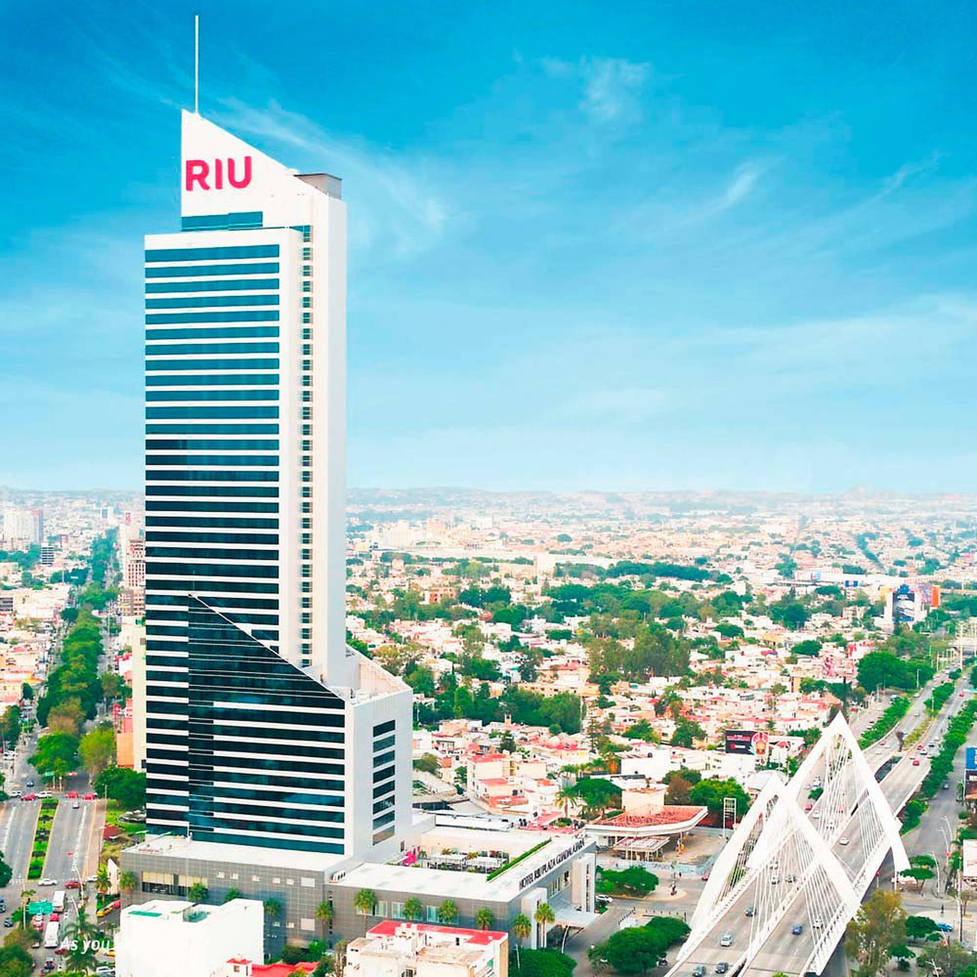 瓜達拉哈拉 Hotel Riu Plaza 壁紙 — 一座高聳的白色RIU酒店摩天大樓在晴朗的藍天和散落的雲朵下主導著充滿活力的城市景觀。