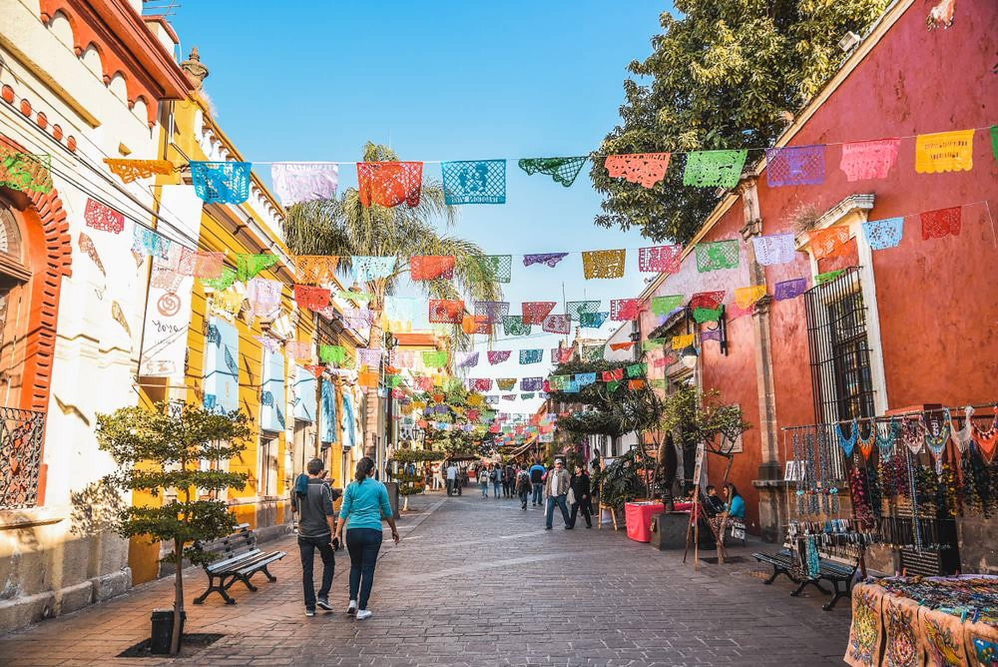 瓜達拉哈拉色彩繽紛的彩旗 壁紙 — 色彩繽紛的 papel picado 橫幅在陽光明媚的鵝卵石街道上飄揚，街道兩旁是色彩繽紛的殖民時期建築和行人。