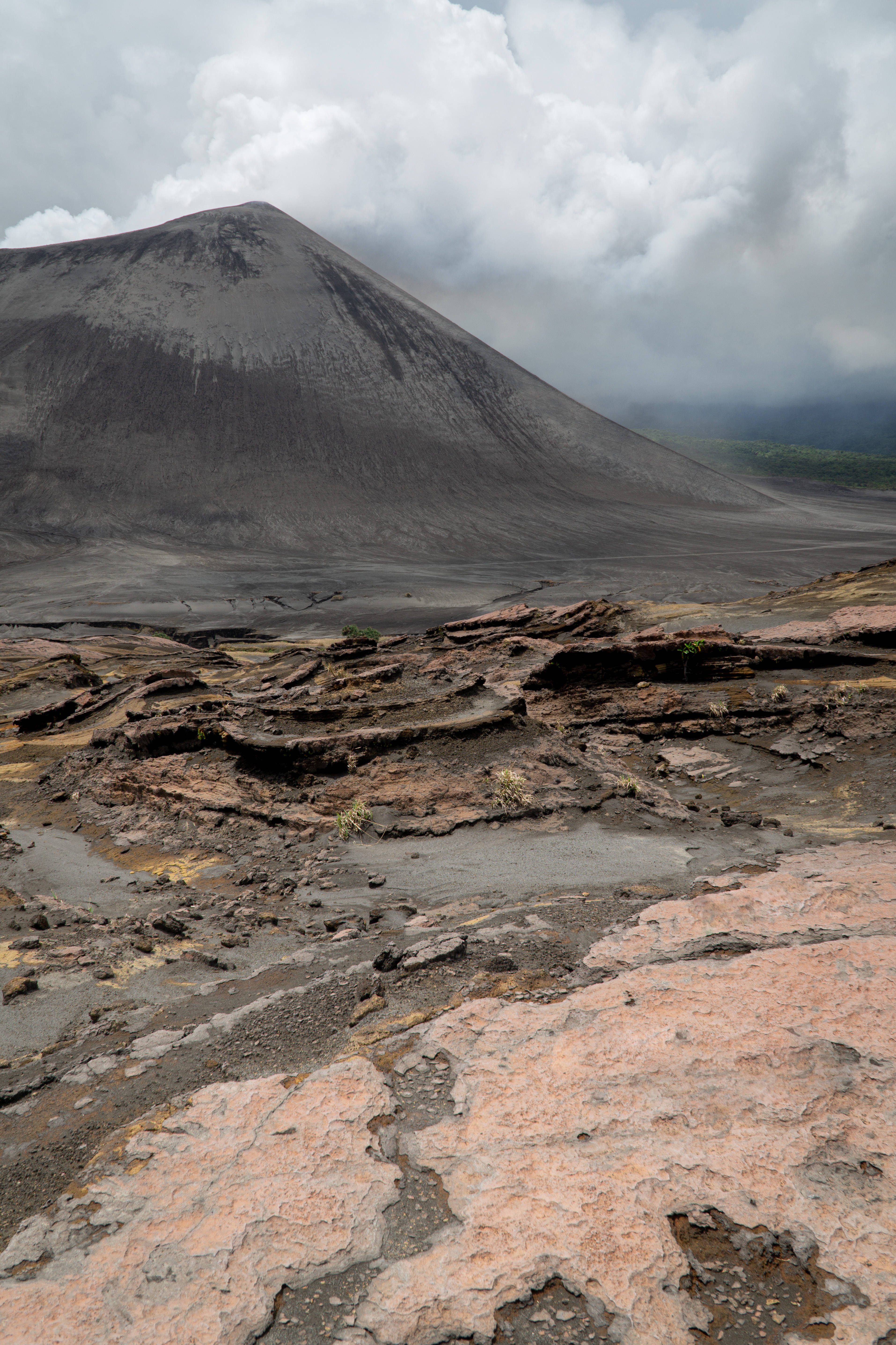 瓦努阿圖灰火山 壁紙 — 一個荒涼的火山景觀，在陰天之下，巨大的灰色火山錐聳立在龜裂的紅褐色地形後方。