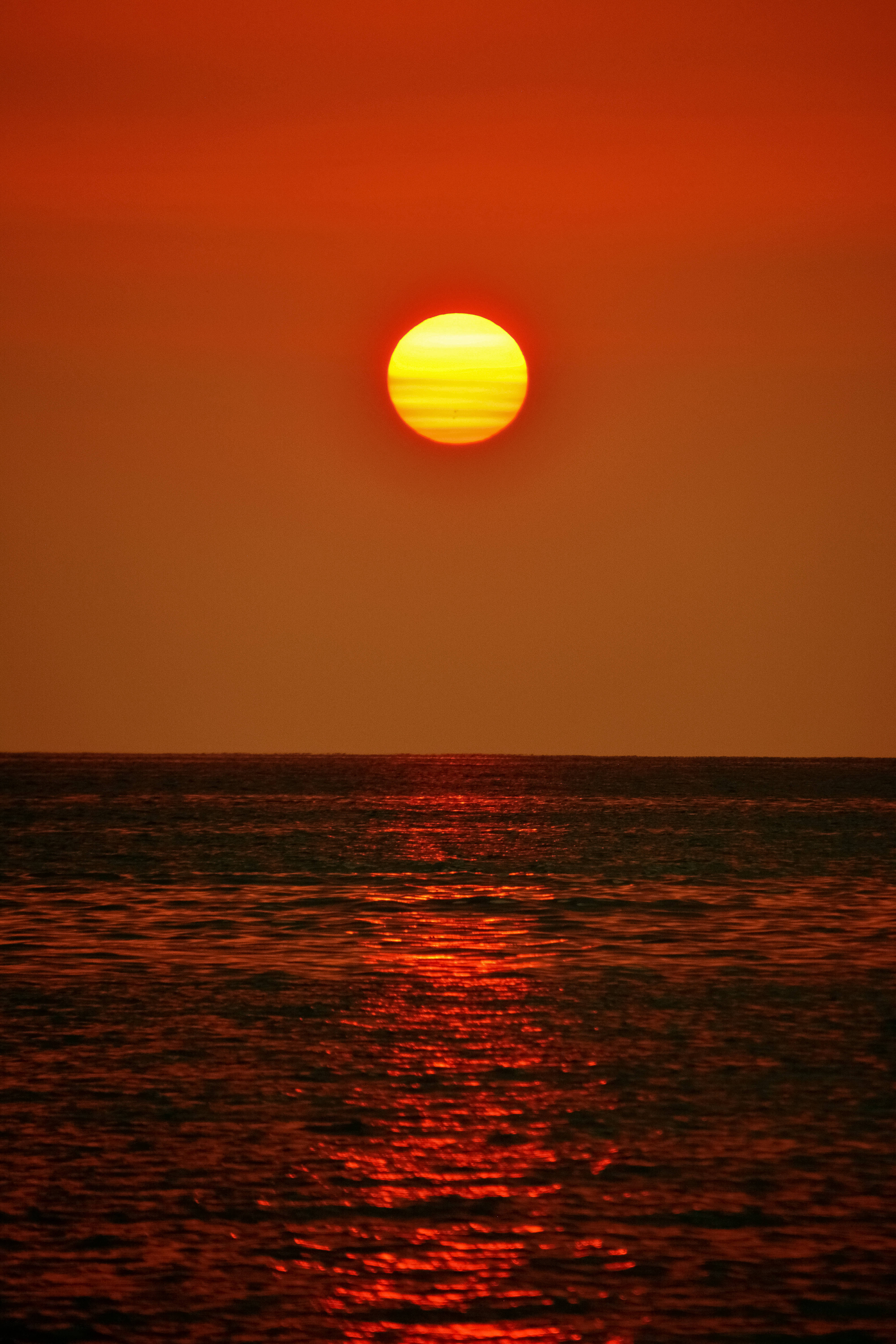 瓦努阿圖紅天夕陽 壁紙 — 一輪熾熱的橙色太陽低掛在平靜的海洋上，其強烈的光芒在漣漪的水面上反射。