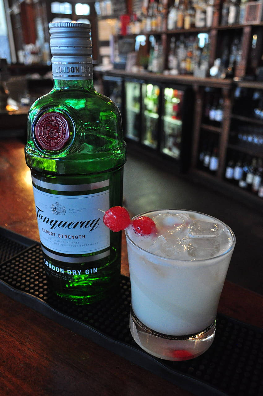 瓶中雅緻——Tanqueray金酒於高級吧檯 壁紙 — 一瓶綠色嘅Tanqueray氈酒放喺吧枱墊上，旁邊係一杯用櫻桃裝飾嘅冰凍雞尾酒。