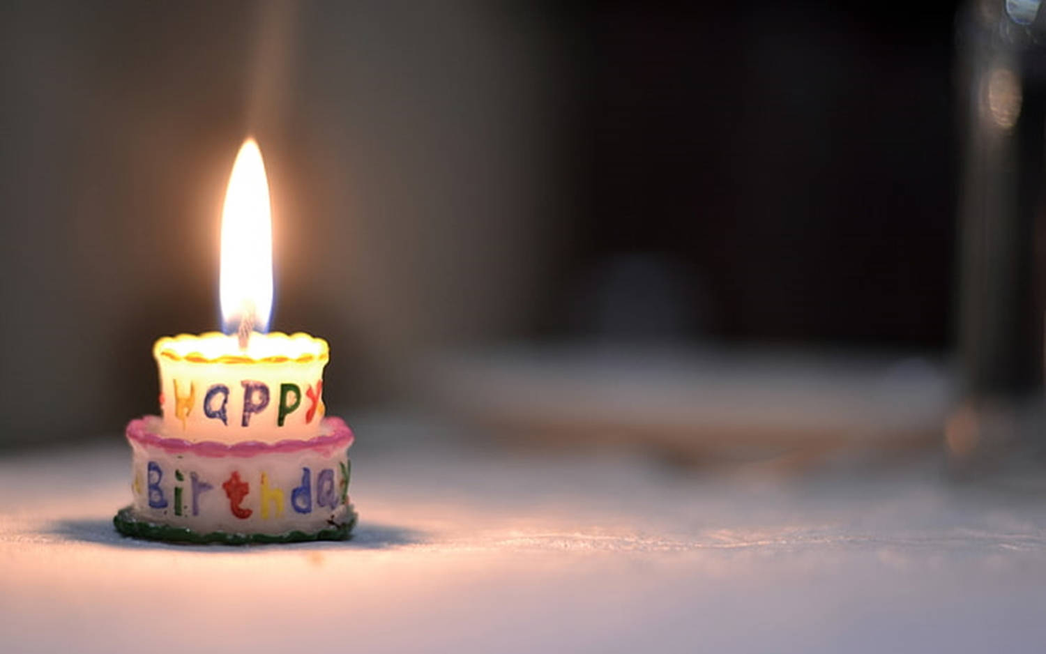 生動慶祝時刻 - 燃亮蠟燭的生日蛋糕 壁紙 — 一個細小的兩層生日蠟燭，帶有彩色字母，在柔和模糊的背景上溫暖地發光。