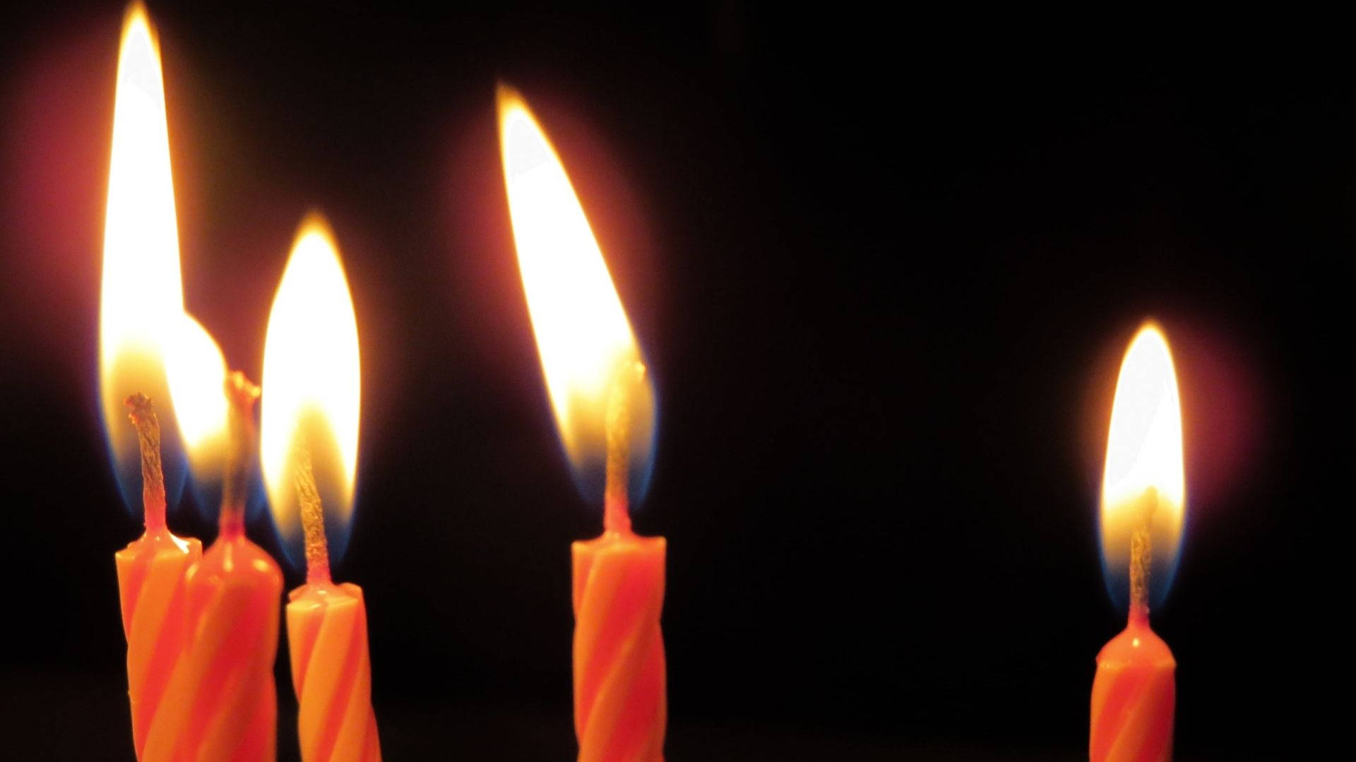 生日蠟燭 壁紙 — 四根橙色扭曲生日蠟燭，帶有明亮的火焰，在黑色背景上溫暖地發光。