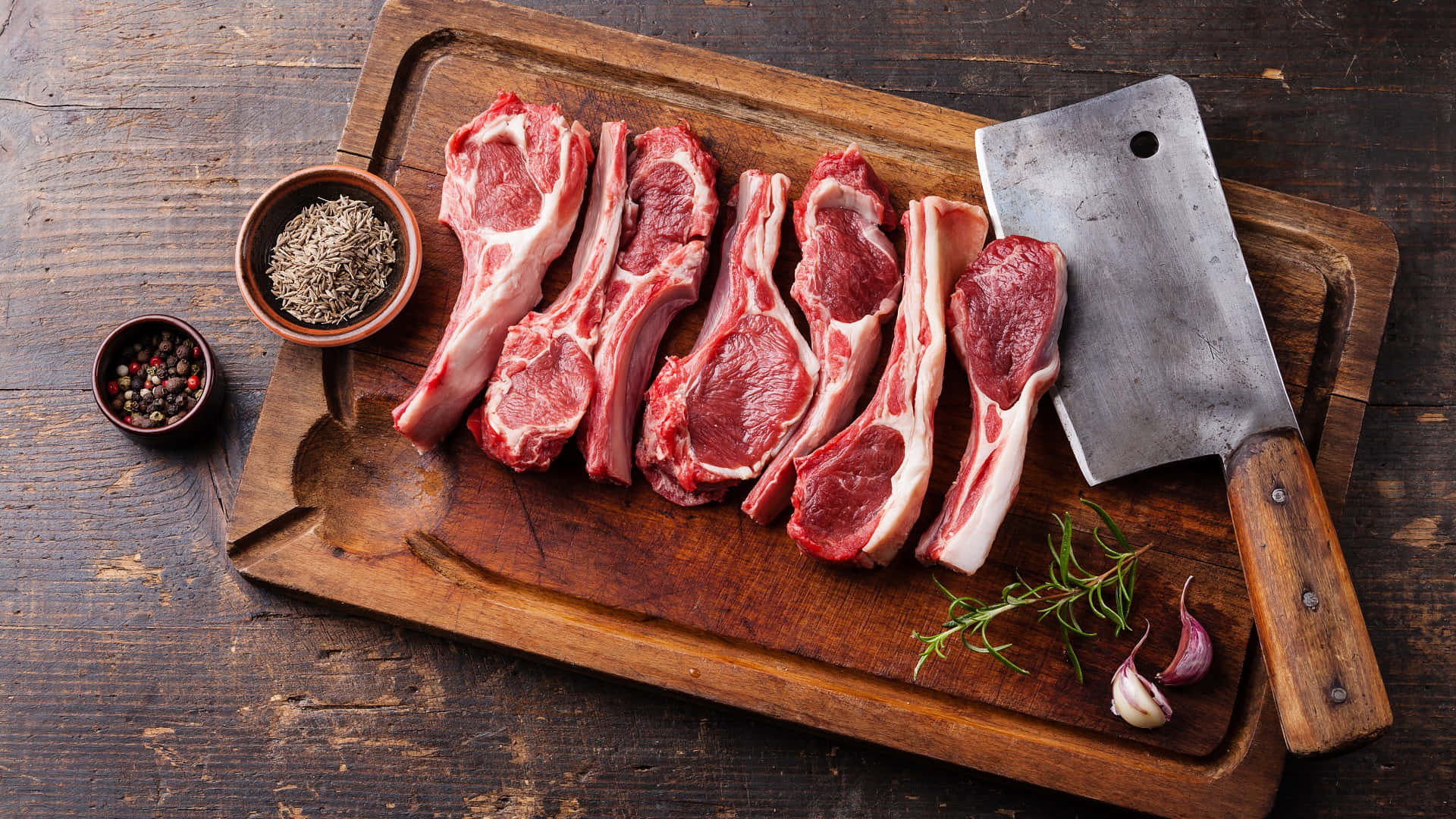 meat 桌布 壁紙 — 生羊扒配迷迭香、大蒜和香料，擺放在質樸的木砧板上，附一把剁肉刀。