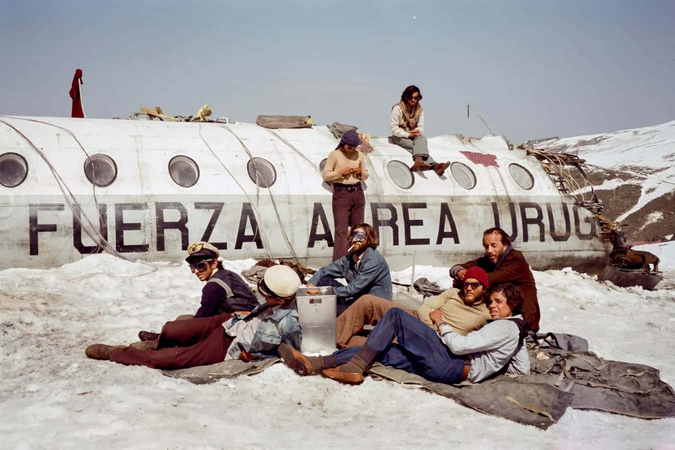  壁紙 — 生還者在荒涼的山區冬季景觀中，於一架失事的烏拉圭軍用飛機旁邊的雪地上放鬆。