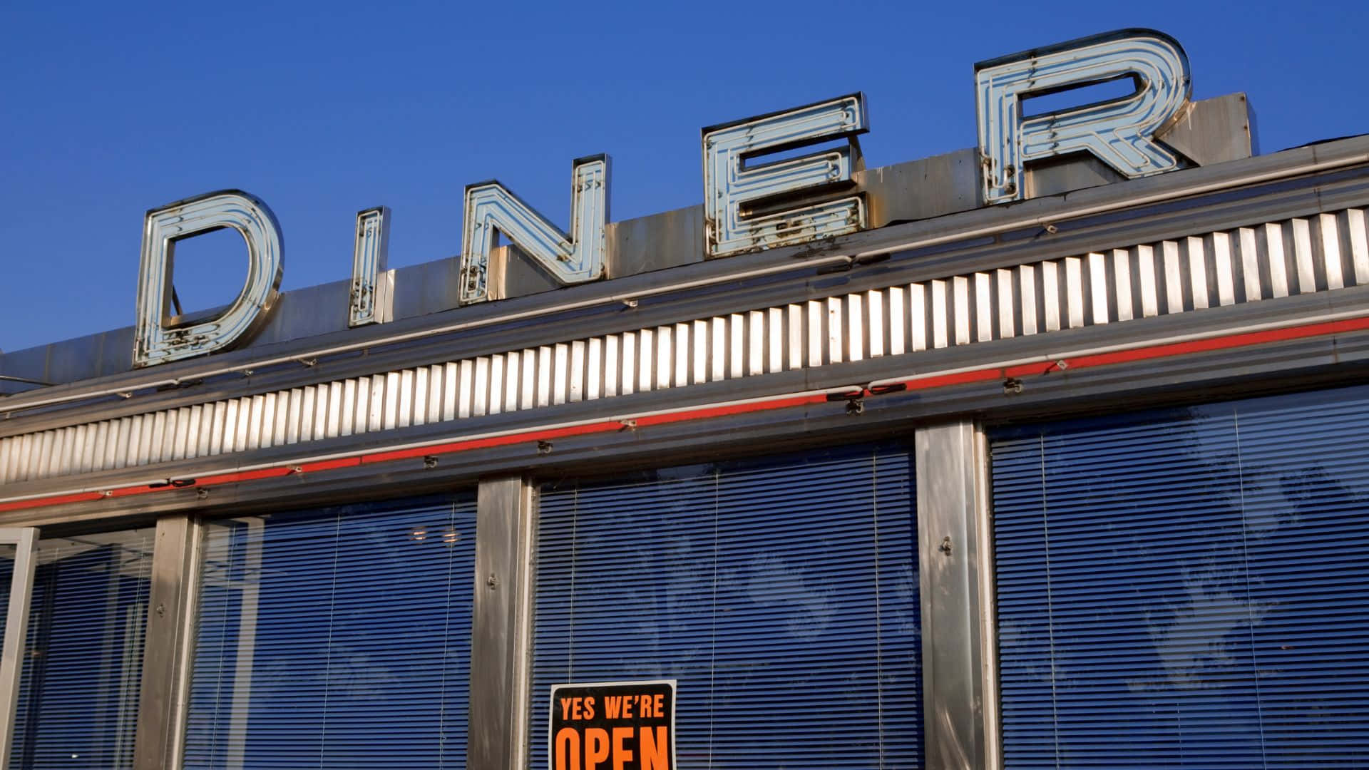 Diner 桌布 壁紙 — 生鏽的霓虹燈 'DINER' 標誌在晴朗的藍天下發光，位於一間拉下百葉窗的復古餐廳上方，下方有一個明亮的 'OPEN' 標誌。