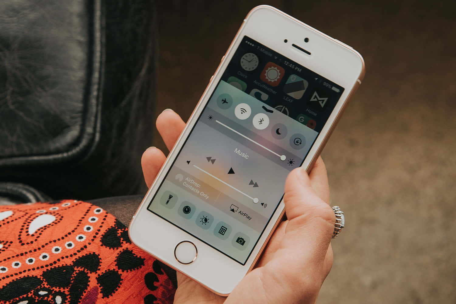 用 Apple 手機捕捉生活精華 壁紙 — 一隻手拿著玫瑰金色iPhone，顯示控制中心，包含音樂播放控制和應用程式圖示，背景為橙色圖案布料。
