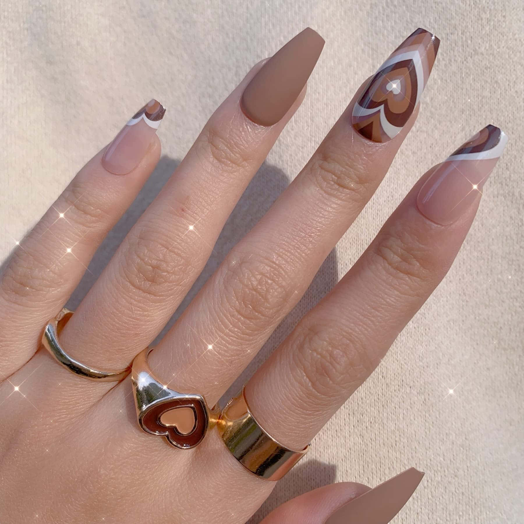 用 Fake Nail 讓你的指甲發揮創意 壁紙 — 一隻手，長有杏仁形指甲，帶有棕色和白色的幾何心形圖案，金色戒指，背景為柔和的米色。