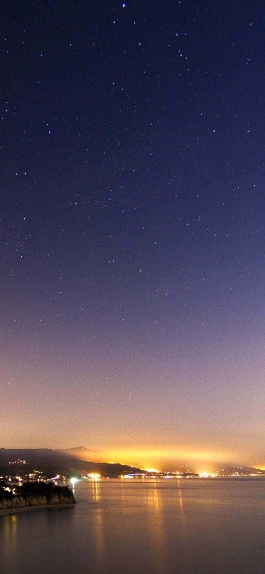 用 iPhone X 捕捉馬利布海灘的絕美風光 壁紙 — 繁星點點的夜空拱形籠罩著平靜的海灣，映照著遠處的城市燈光和發光的山丘。