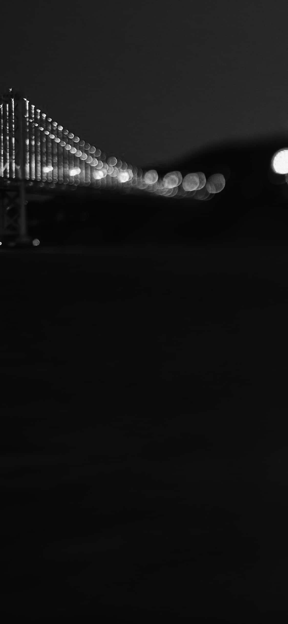 用 iPhone X 欣賞加州海灘美景 壁紙 — 一張黑白散景照片，拍攝夜晚中燈火通明的吊橋，發光球體在黑暗天空的襯托下。