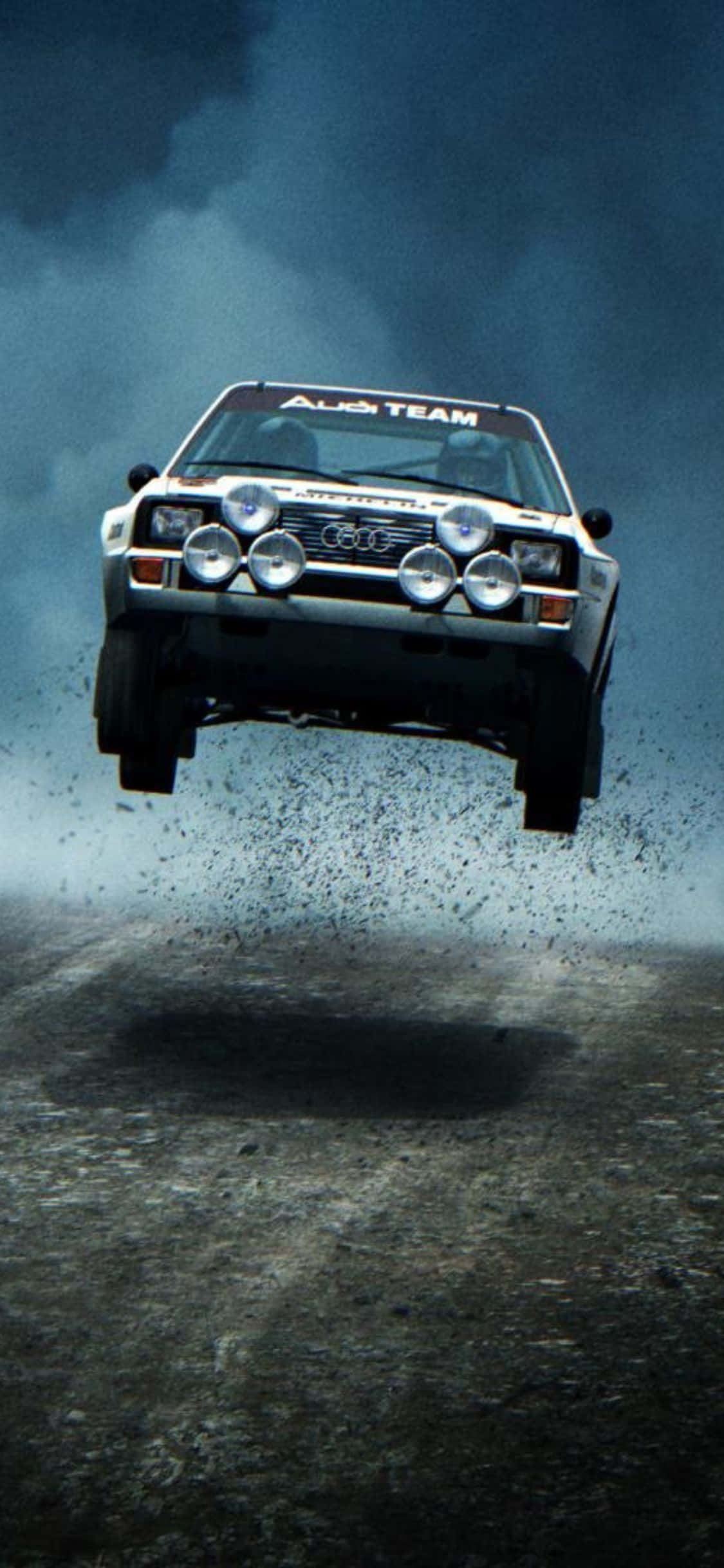 用 iPhone Xs Dirt Rally 感受賽車的興奮 壁紙 — 一輛白色奧迪拉力賽車在暴風雨的天空下，在泥濘賽道上騰空而起，揚起碎石。