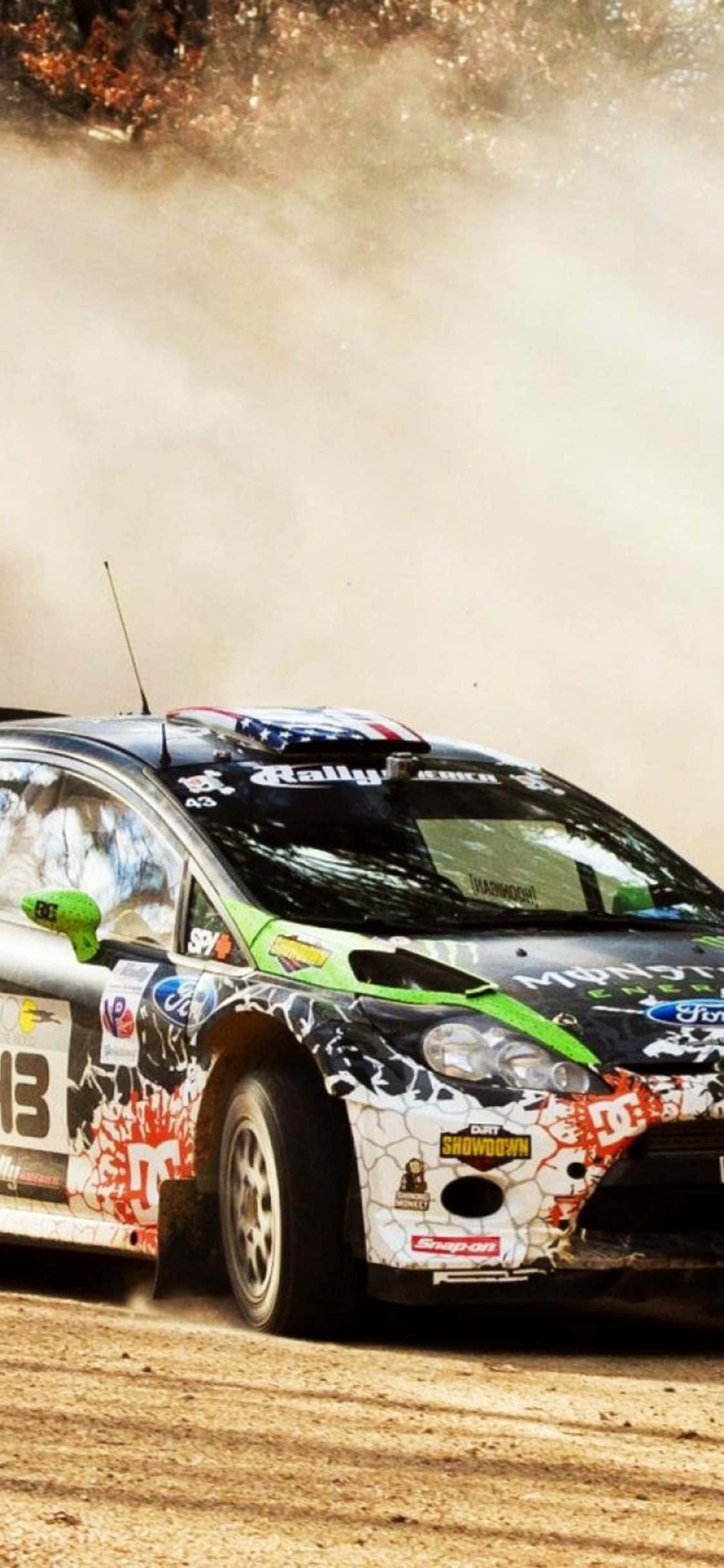 用 iPhone Xs 和 Dirt Rally 將你的賽車夢想推向新境界 壁紙 — 一輛拉力賽車在崎嶇的泥濘賽道上高速飛馳，揚起塵土，上演一場高辛烷值的比賽。
