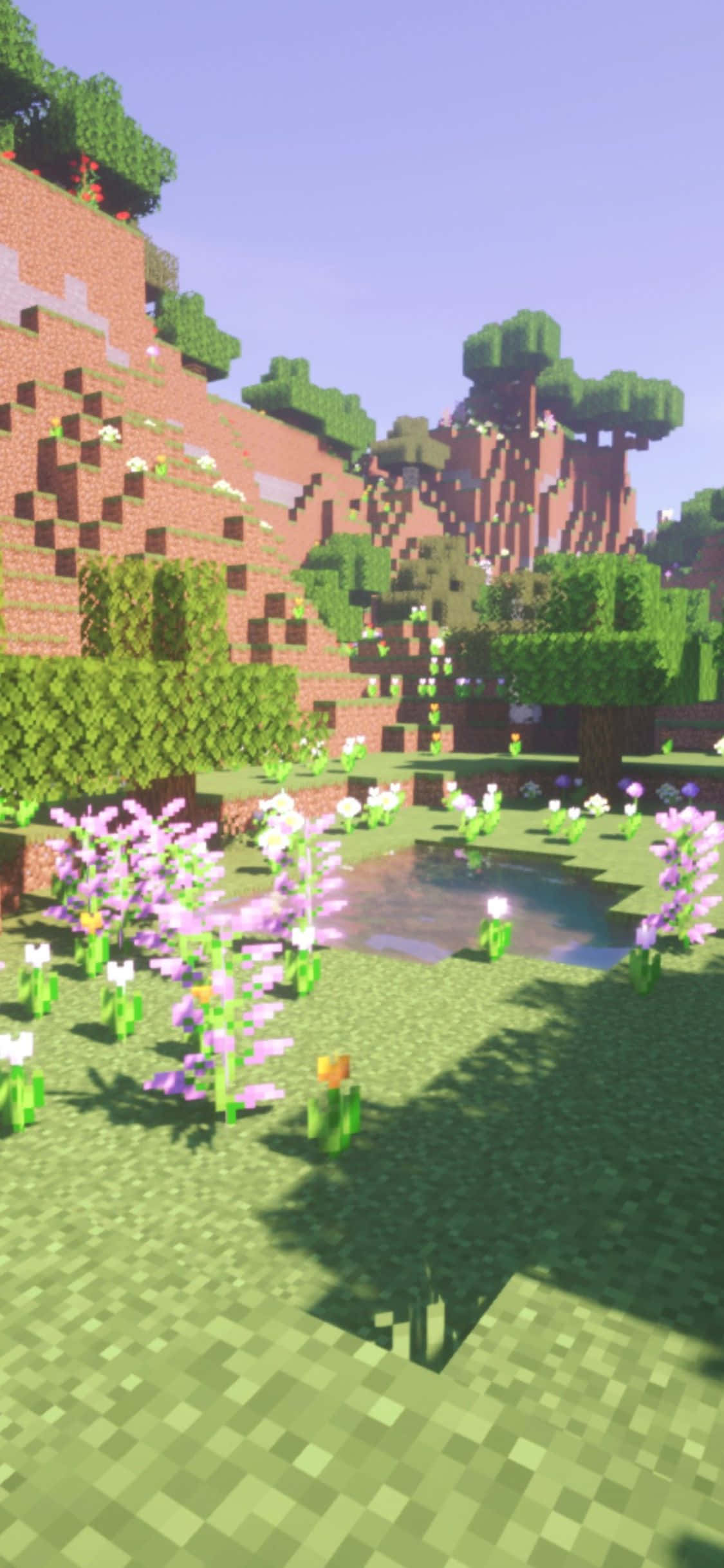 用 iPhone Xs 在 Minecraft 中創造無限可能 壁紙 — 一個像素化的Minecraft景觀，有綠色草地、紫色花朵和一個被方塊狀樹木和山丘環繞的小池塘，天空晴朗。