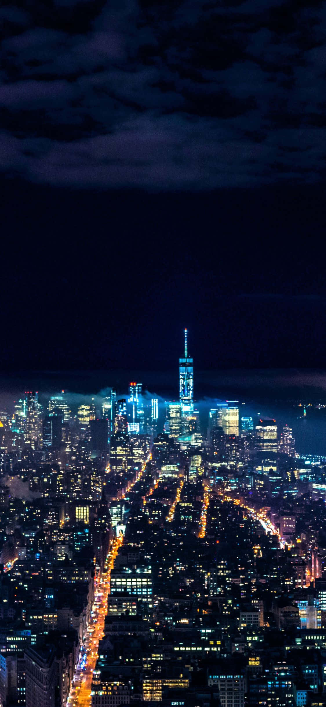 用 iPhone Xs 感受紐約市中心的活力 壁紙 — 紐約市天際線的夜間空中景觀，在陰暗多雲的天空下，摩天大樓燈火通明，街道閃爍著光芒。