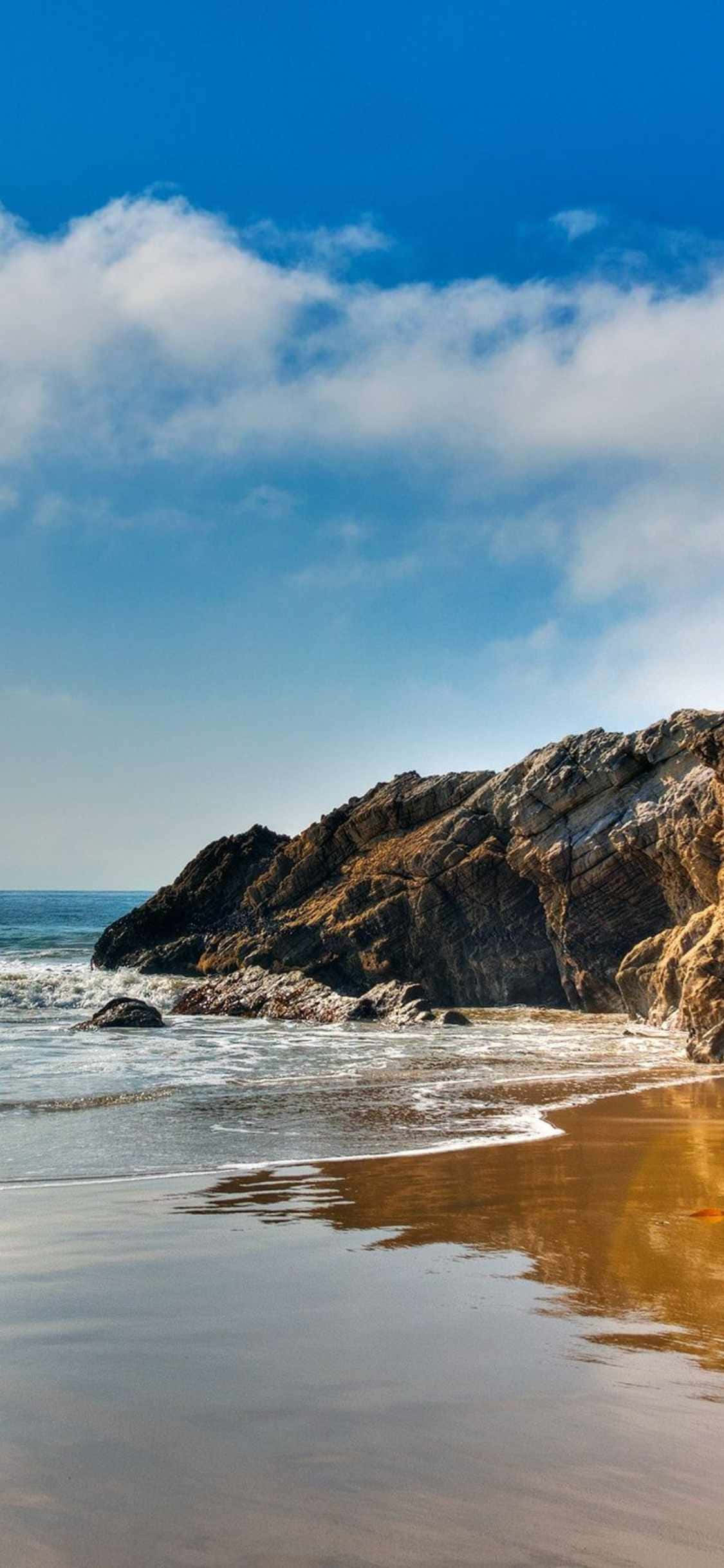 用 iPhone Xs 馬利布欣賞壯麗海景 壁紙 — 陽光明媚的海灘，金色沙灘和拍打崎嶇懸崖的海浪，在明亮的藍天下有蓬鬆的白雲。