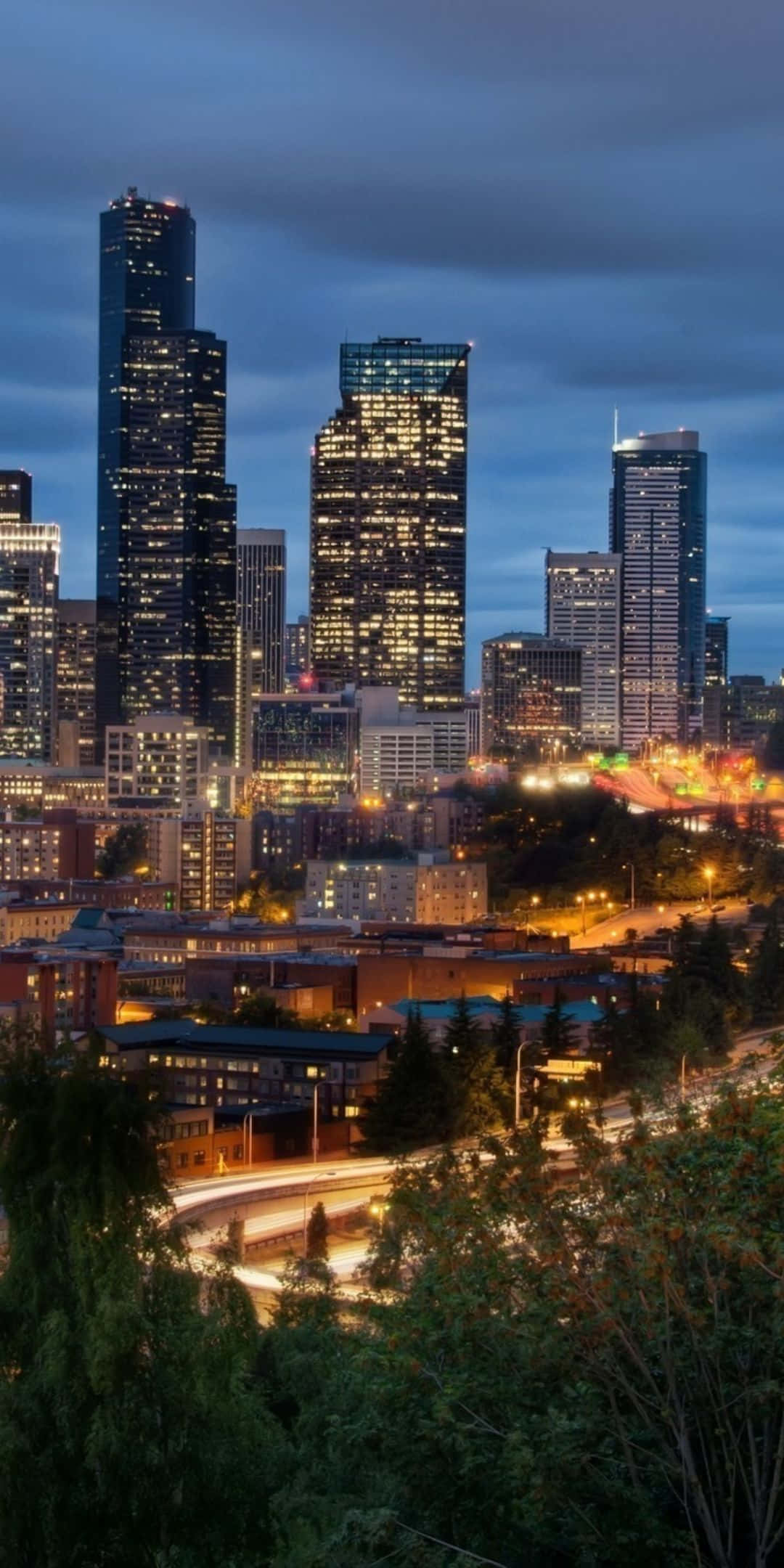 用 Pixel 3 探索西雅圖天際線 壁紙 — 暮色城市景觀，高聳的摩天大樓在昏暗的天空下閃耀，周圍環繞著茂密的綠樹。