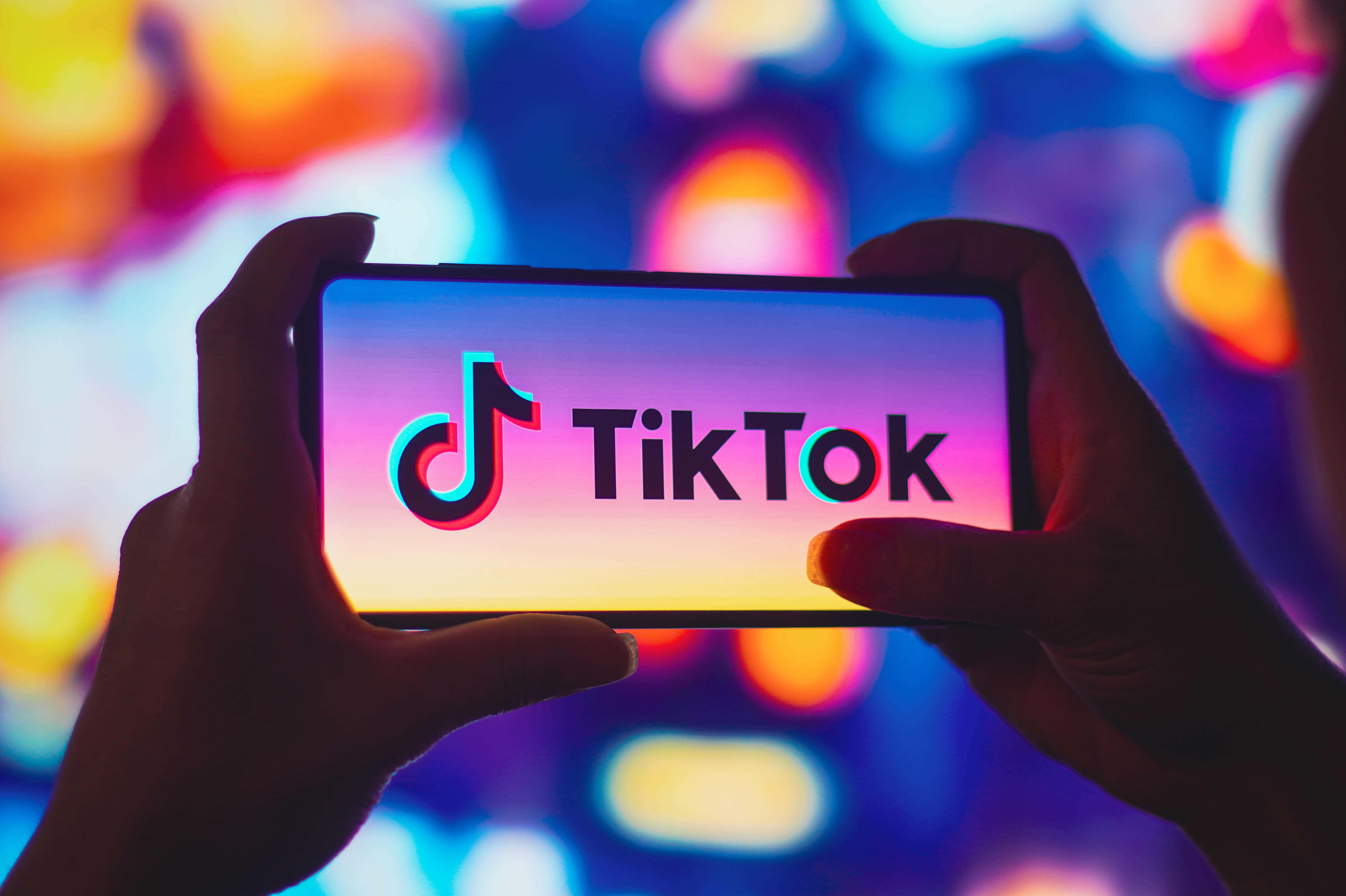用 TikTok 表達自我 壁紙 — 雙手拿著一部顯示TikTok標誌的智能手機，背景是色彩繽紛的燈光散景。