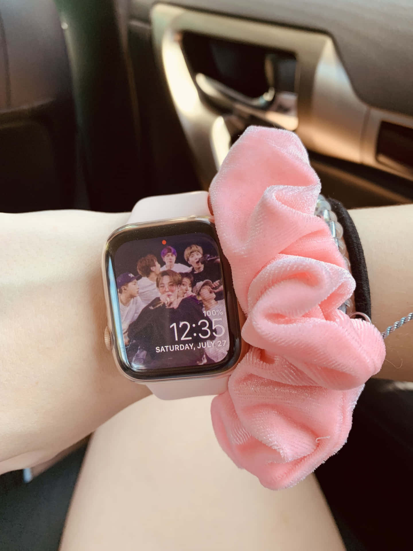 手腕戴粉紅色髮圈和智能手錶，顯示車內K-pop組合照片。