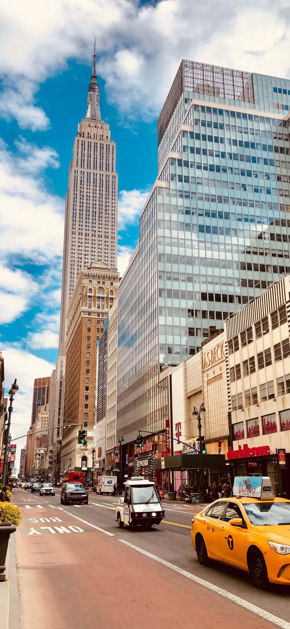 用iPhone XS體驗紐約市的絕美風光 壁紙 — 充滿活力的紐約市街景，黃色出租車和帝國大廈在明亮的藍天和白雲下高聳入雲。