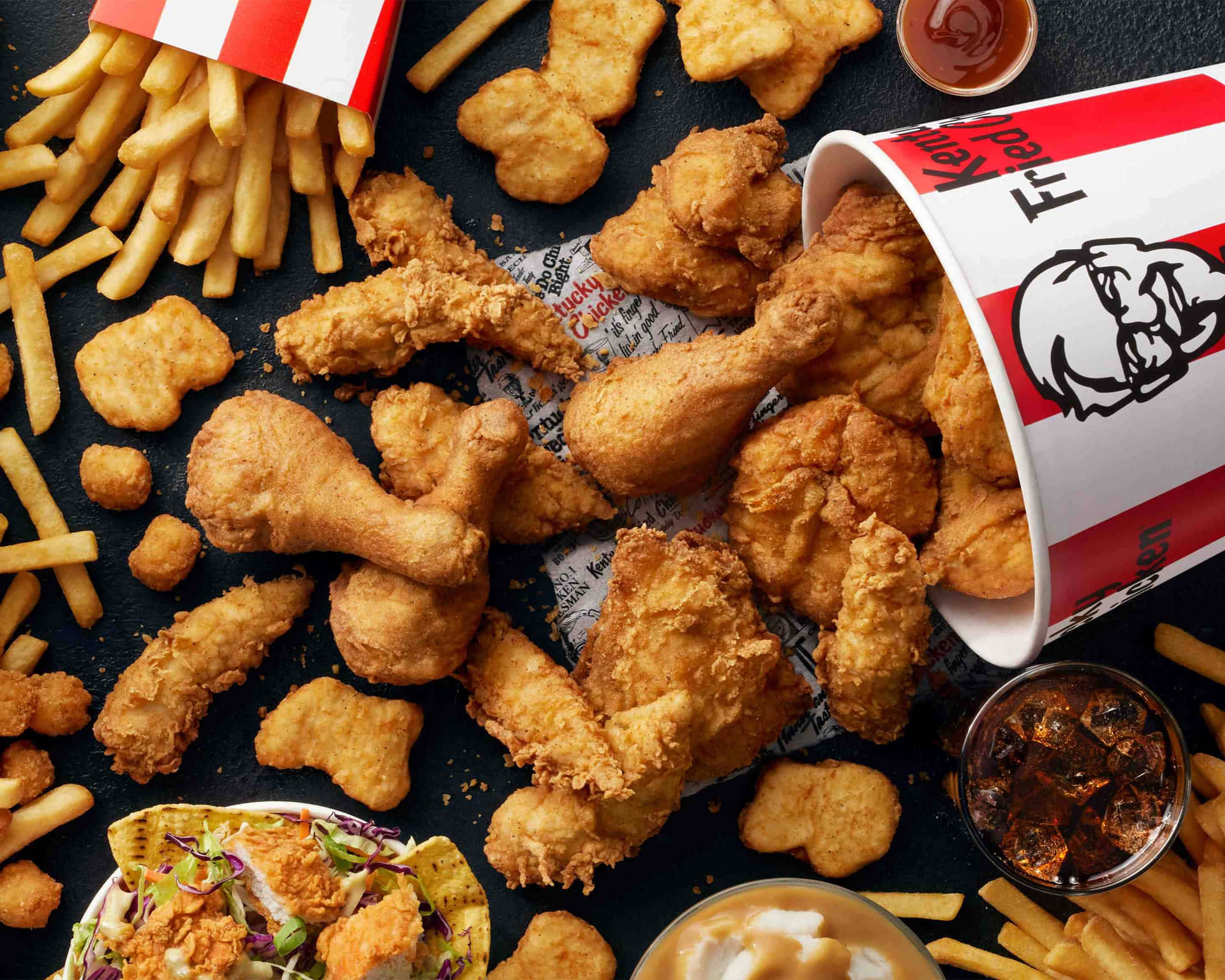 從KFC桶中灑落在深色表面上的炸雞、薯條和醬料。