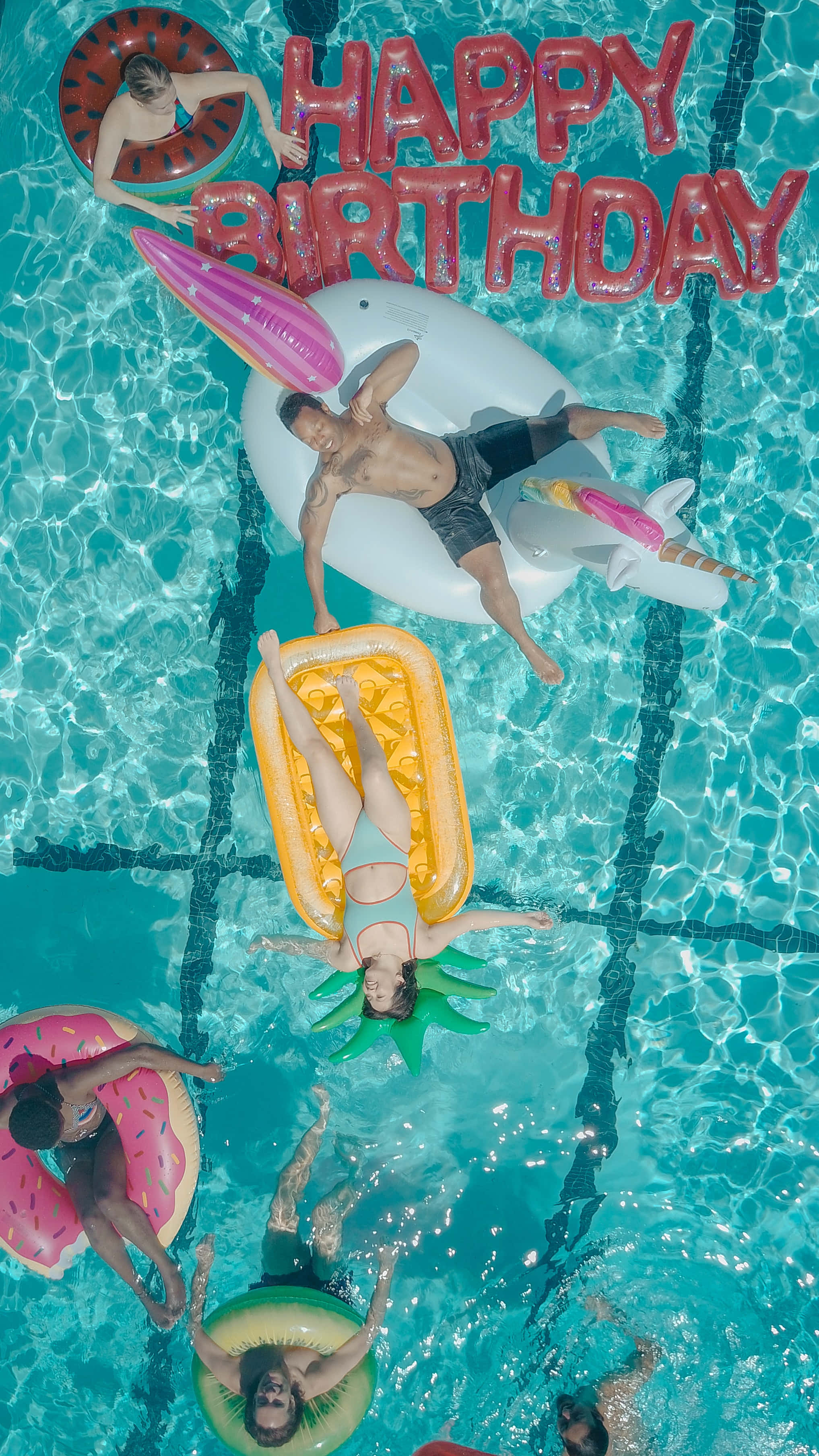 用可愛甜美的生日慶祝特別的一天 壁紙 — Top-down view of friends floating on colorful inflatable toys in a bright turquoise pool with a large HAPPY BIRTHDAY balloon.