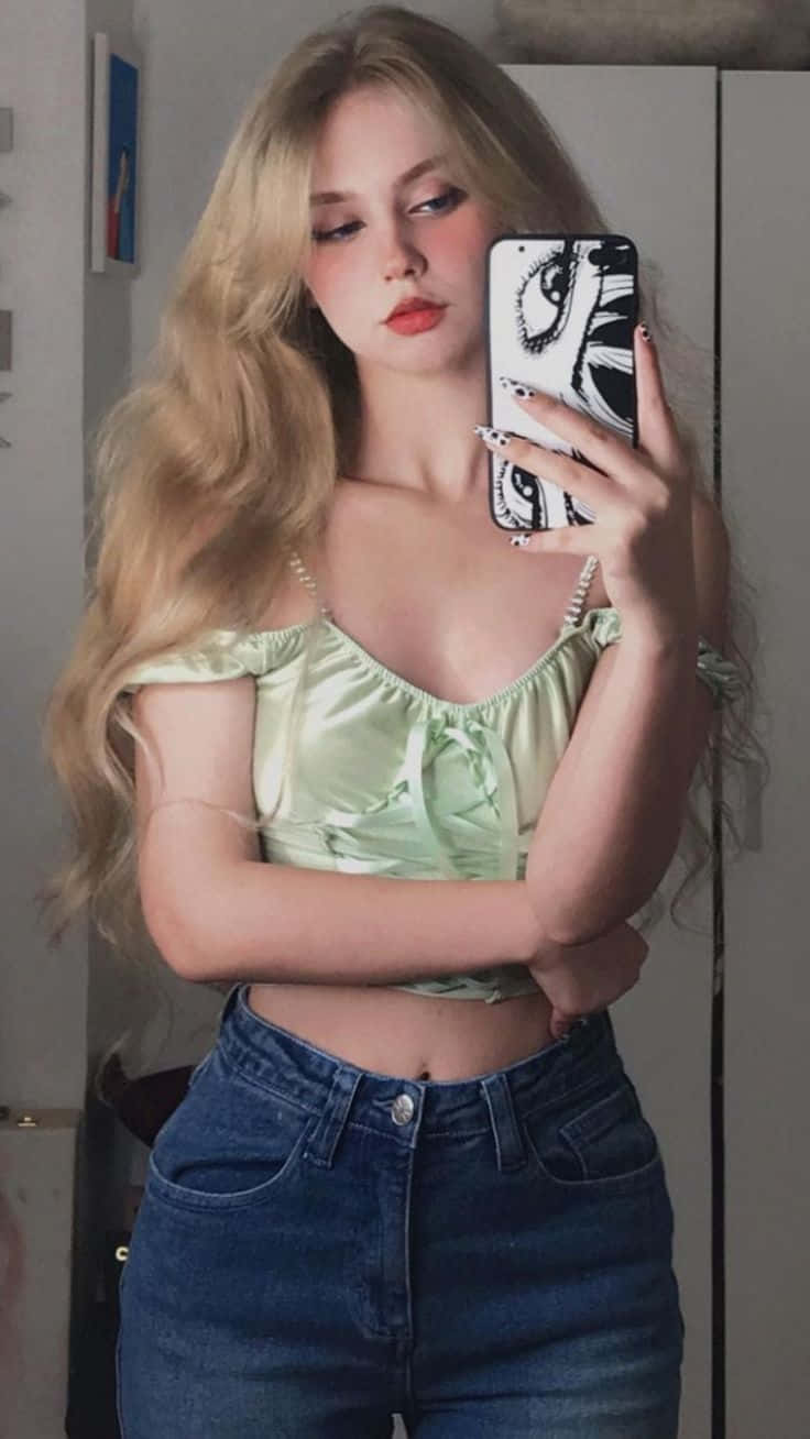 長金髮的年輕女性穿著綠色上衣和牛仔褲對著鏡子自拍。