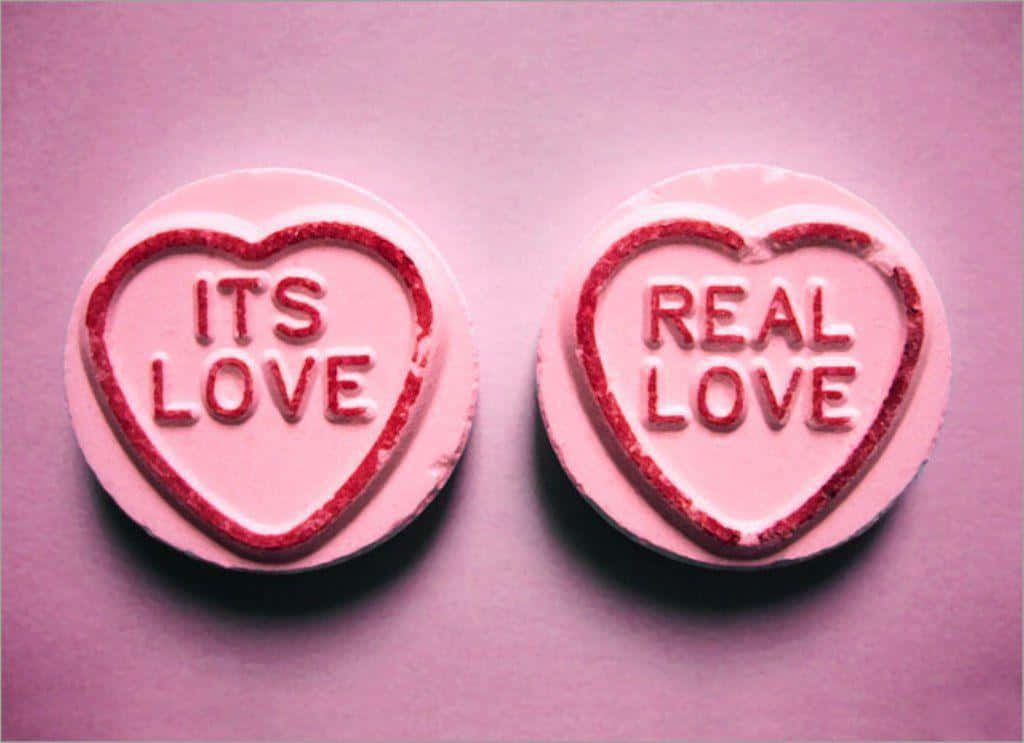 兩粒粉紅色心形糖果，帶有紅色閃粉文字，上面寫著「ITS LOVE」和「REAL LOVE」。