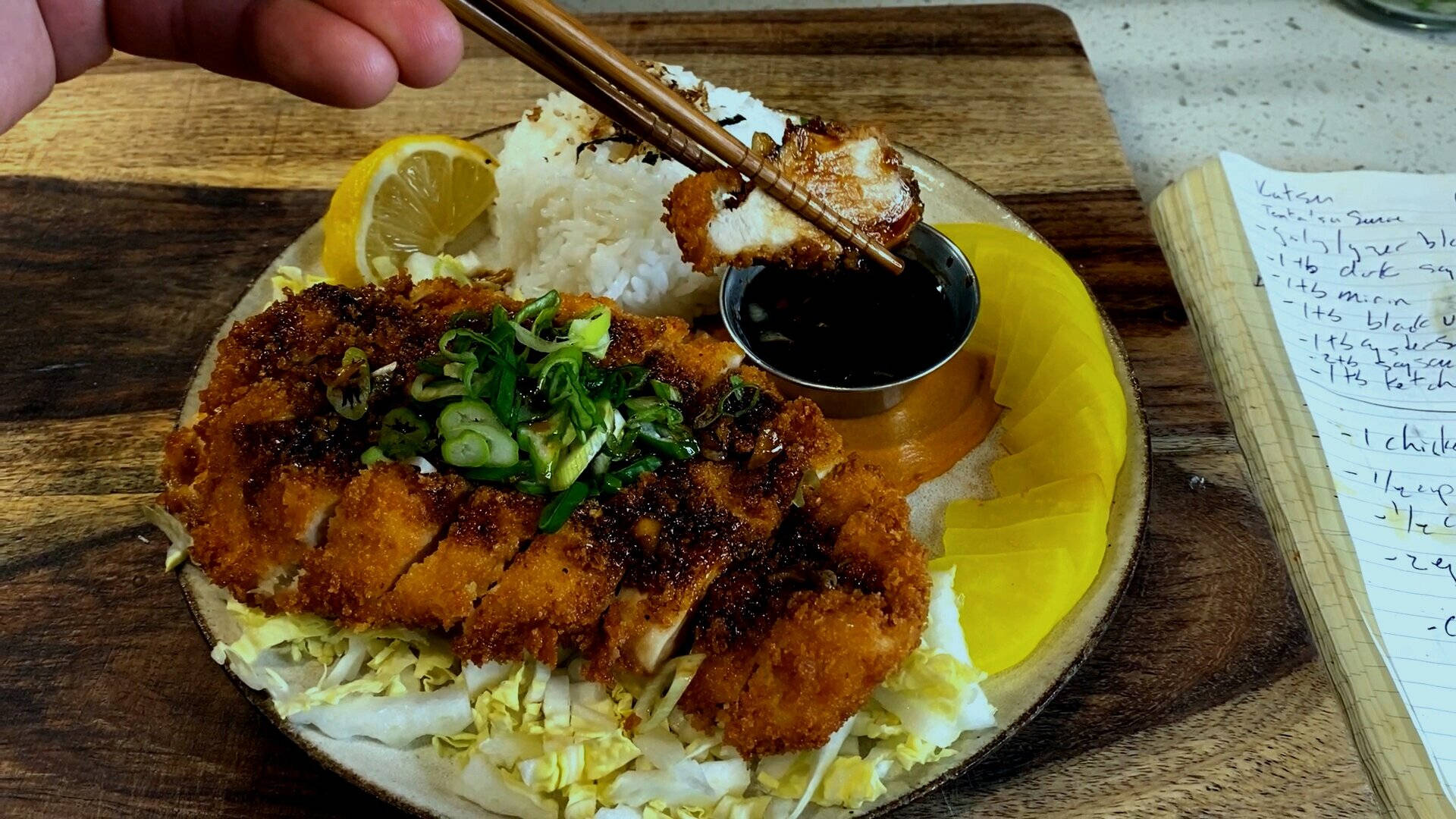 用手筷夾起豬排 壁紙 — 一隻手用筷子從鋪有米飯、椰菜和檸檬角的碟上夾起酥脆的炸豬排。