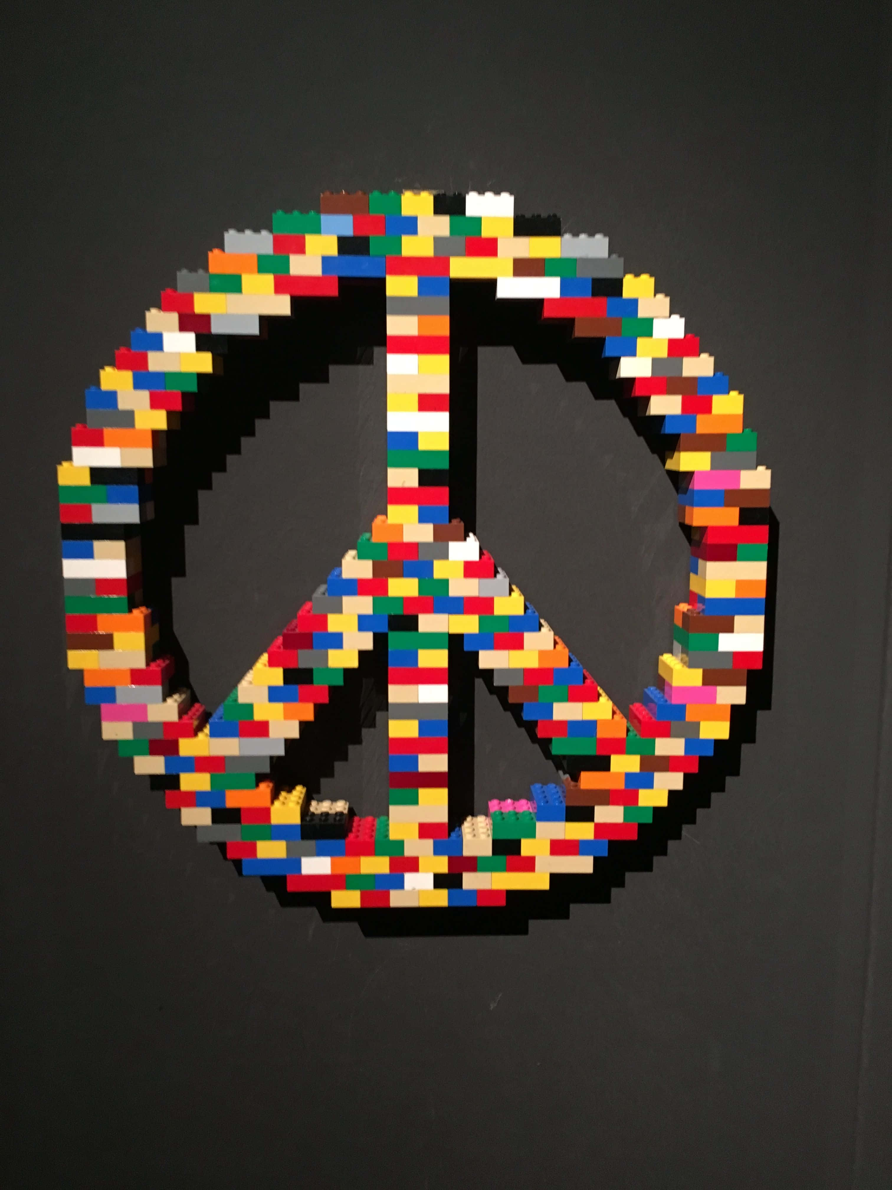 用樂高積木創造驚人藝術作品！ 壁紙 — 一個由多色LEGO積木製成的鮮明和平標誌，在深灰色牆壁上格外突出。