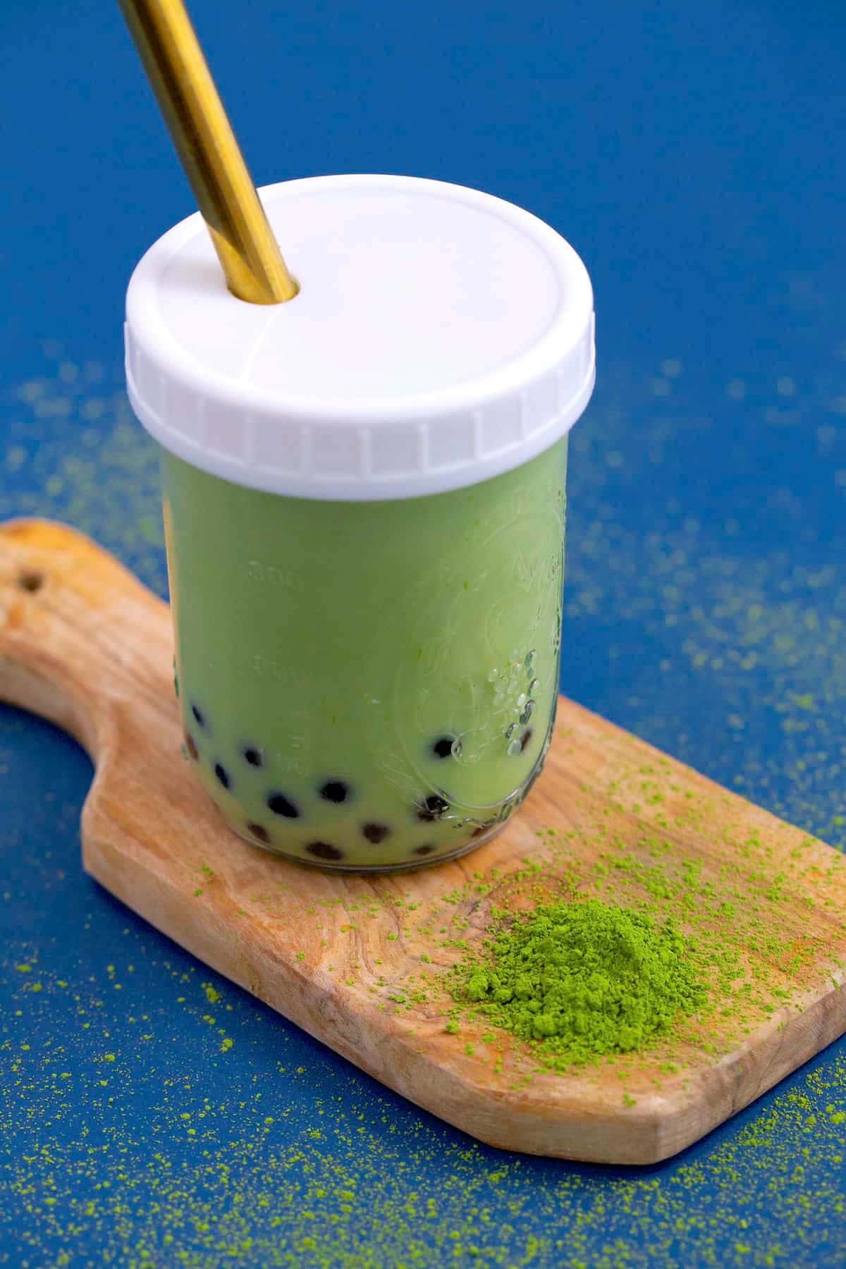用珍珠奶茶為一天注入活力 壁紙 — 一杯充滿活力的綠色抹茶泡泡茶，放在透明杯中，配金色吸管，置於木板上，背景為藍色。