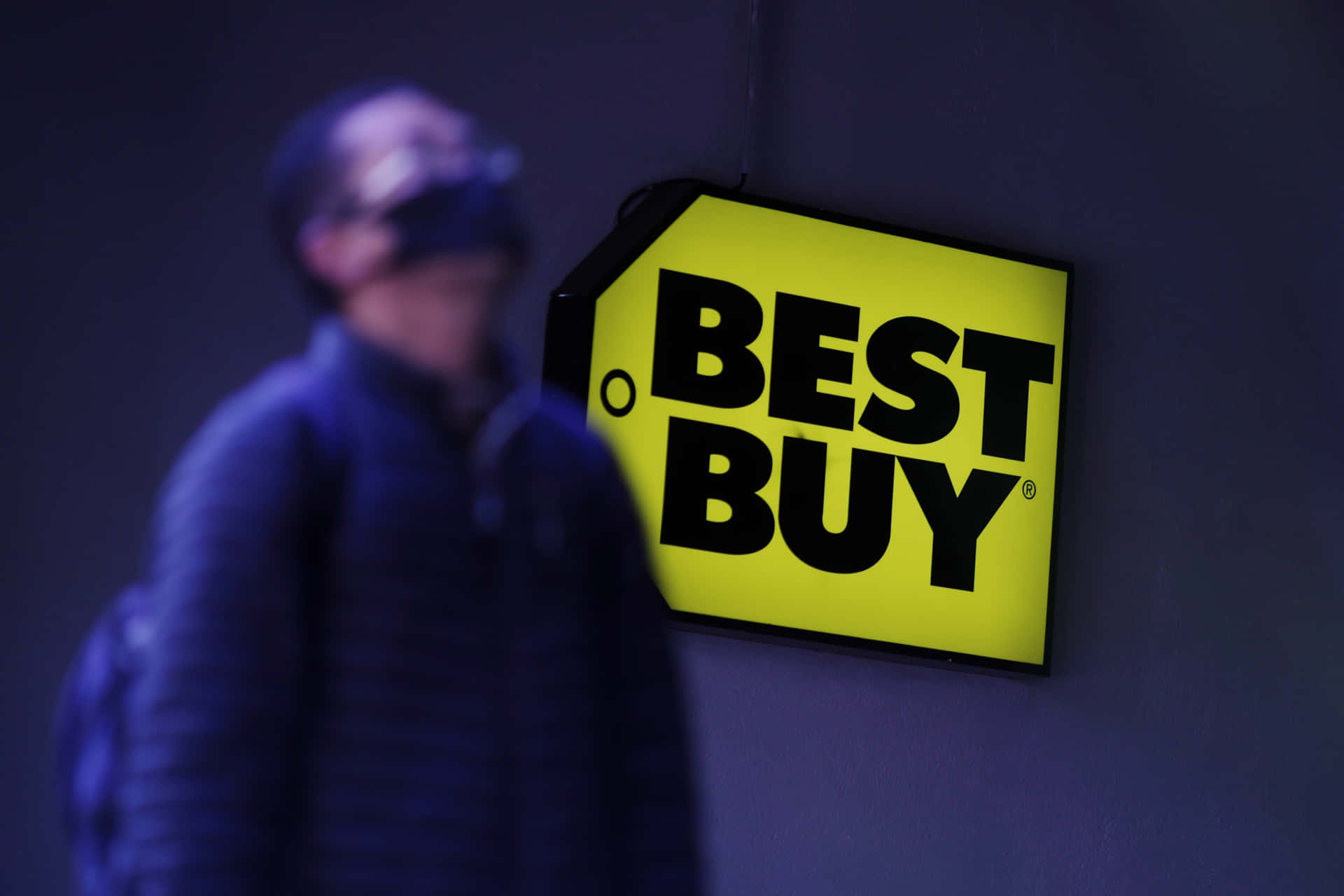 一個發光的Best Buy標誌，在昏暗的城市環境中有一個模糊的人影走過。