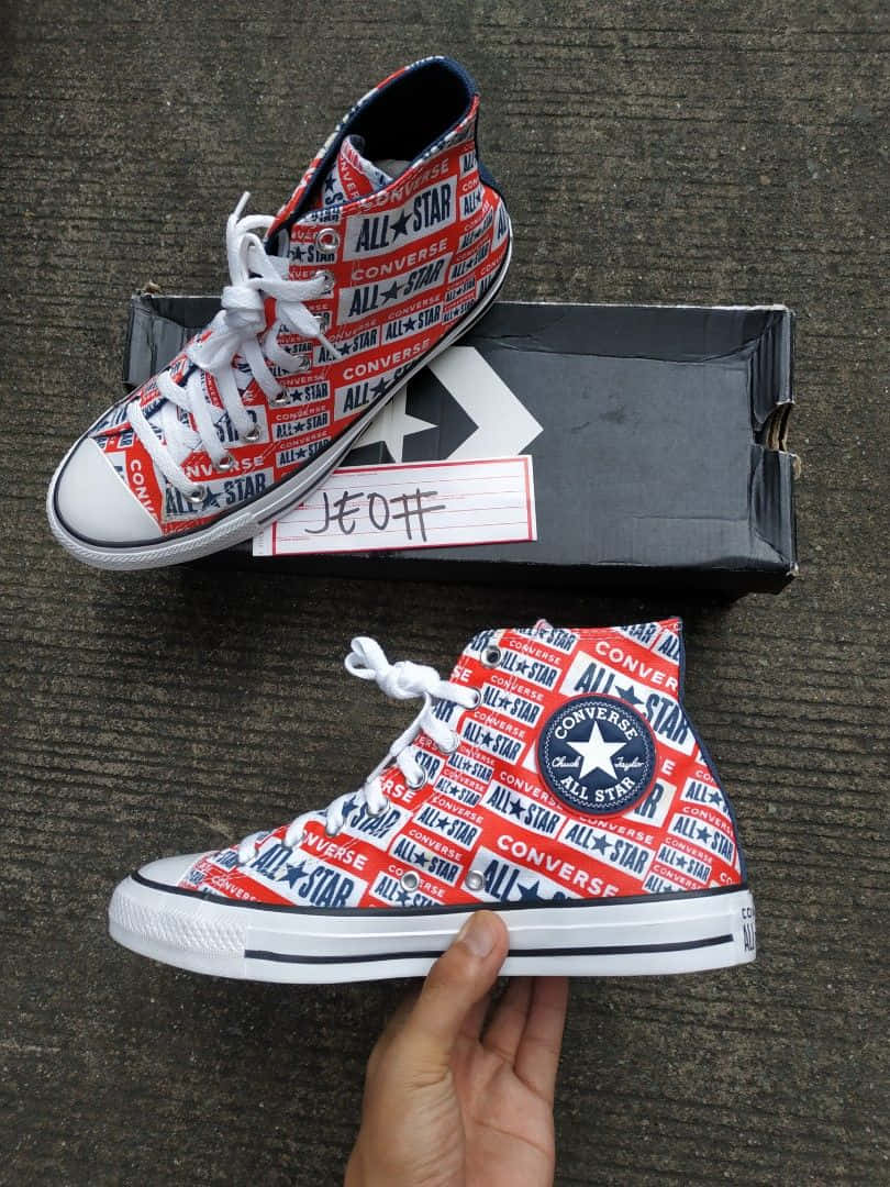 「用經典匡威鞋款製造風格宣言」 壁紙 — 一隻手拿著紅白藍色的Converse All Star高筒運動鞋，鞋上重複印有品牌標誌，背景是紋理灰色地板。