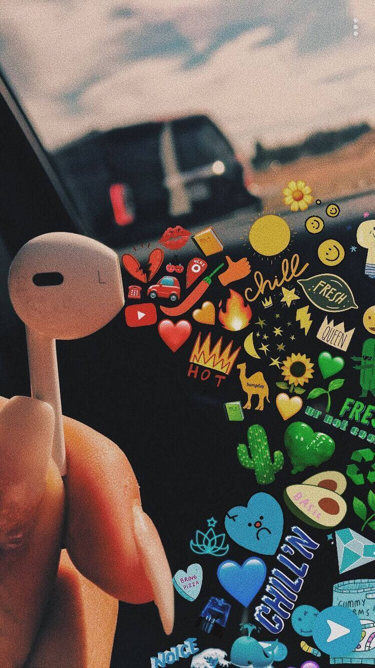 用這些時尚 Airpods 聽你最愛的音樂 壁紙 — 一隻手拿著一個白色耳塞，其屏幕上爆出色彩繽紛的卡通貼紙，背景是模糊的車窗。