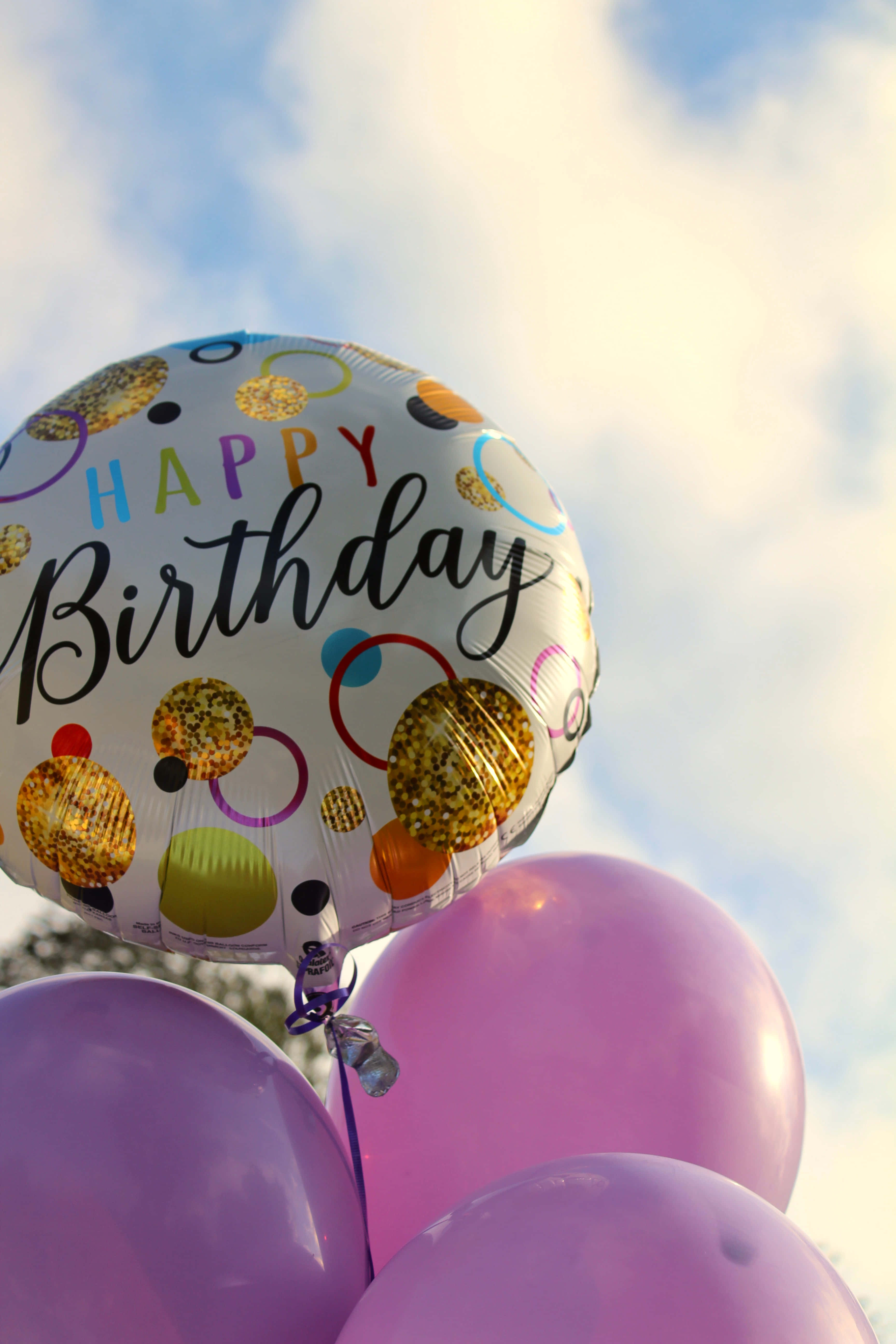 用這款色彩繽紛的生日背景慶祝特別的日子 壁紙 — 一個節日氣氛的 'Happy Birthday' 箔氣球，帶有金色紙屑和彩色圓圈，在明亮的天空背景下漂浮在紫色氣球上方。