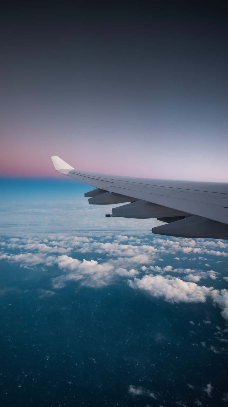 用飛機造型iPhone記錄你的旅程 壁紙 — 飛機機翼延伸至蓬鬆的白雲之上，背景是帶有柔和粉紅色和藍色色調的暮色天空。