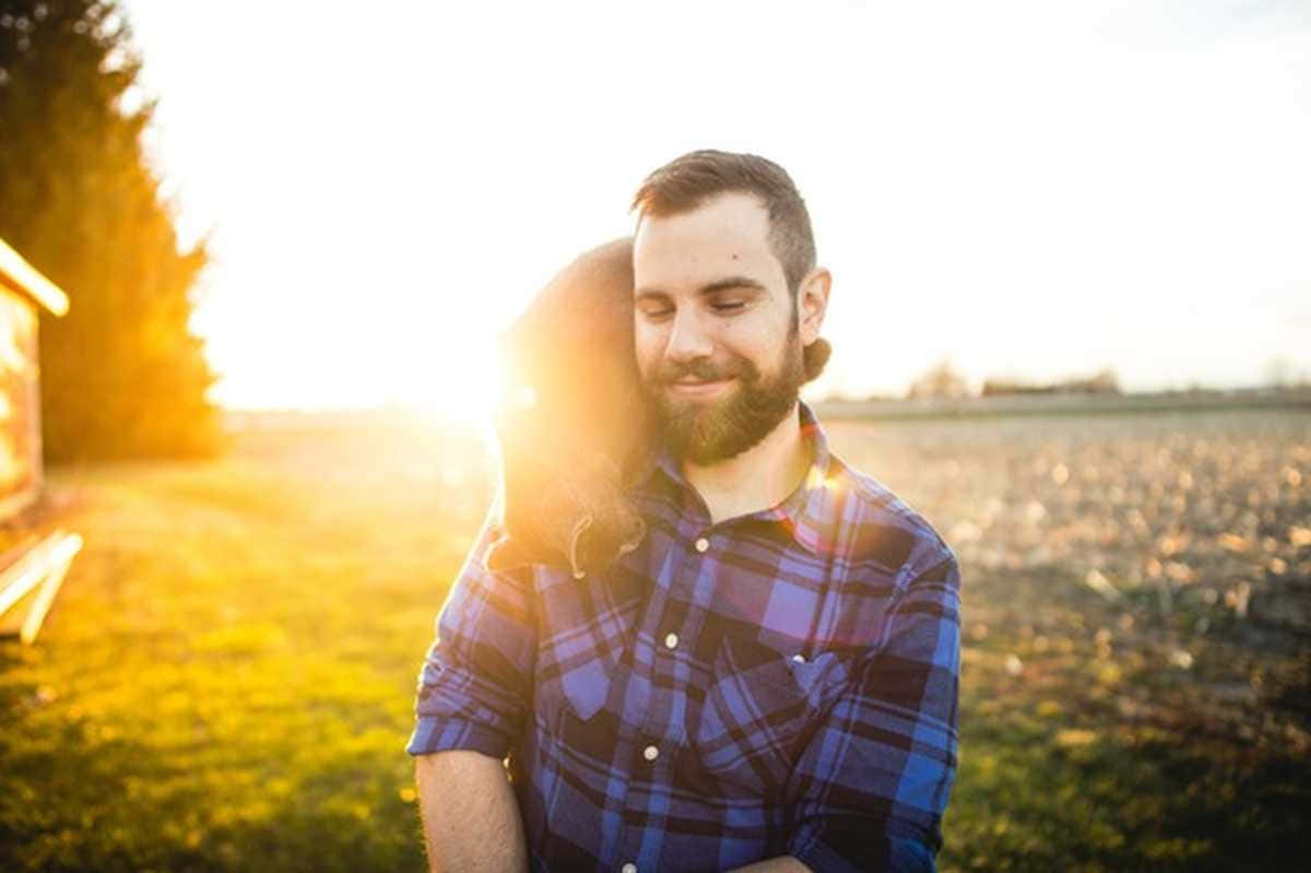男人與寵物夕陽側影照 壁紙 — 一個留鬍鬚的男人穿著格仔恤衫擁抱一個女人，金色夕陽的光線在他們身後的草地上閃耀。