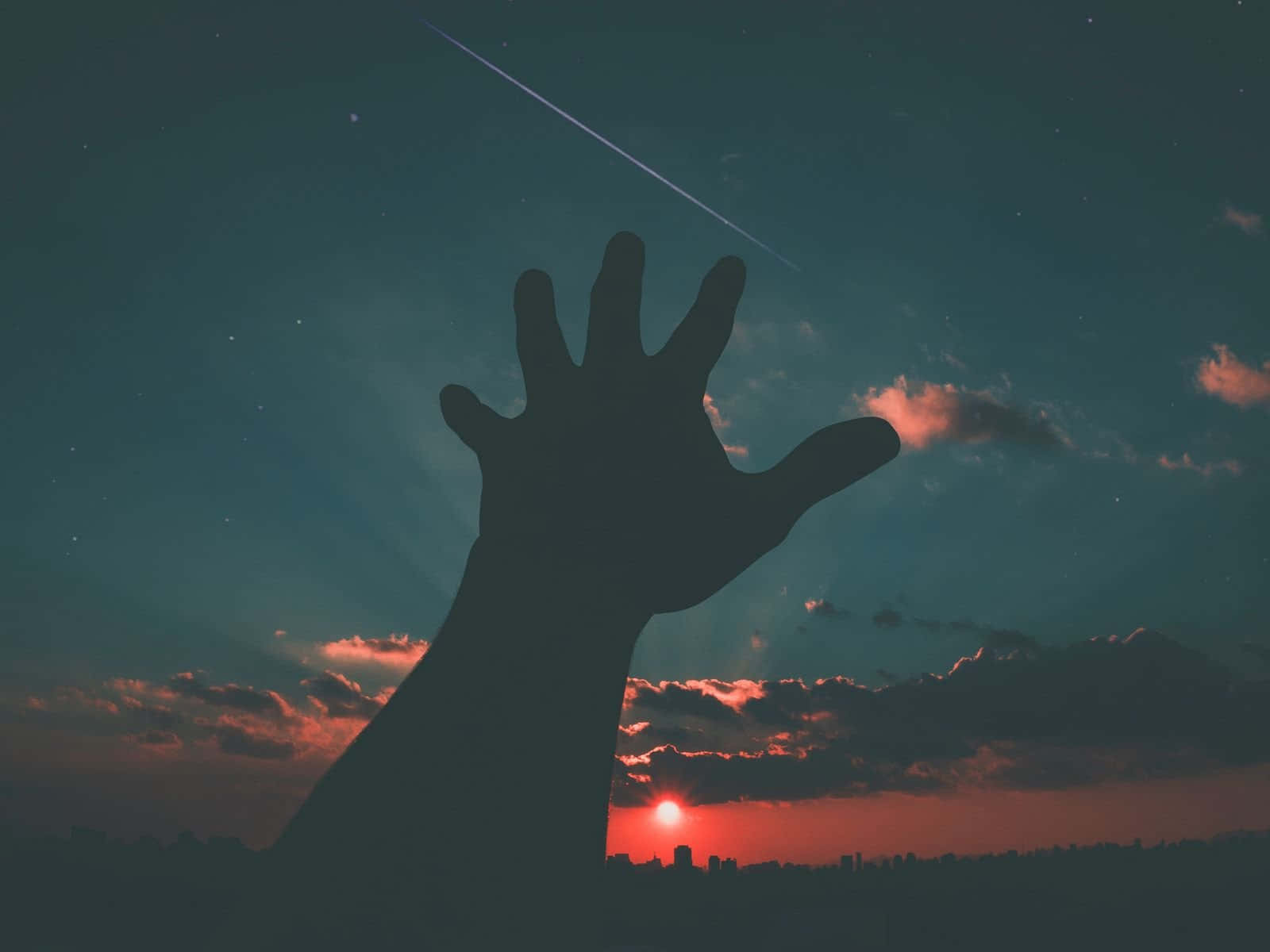 男子伸手向夜空月圖 壁紙 — 一隻剪影手向上伸展，對著佈滿星星和發光日落的暮色天空，背景是遙遠的城市景觀。