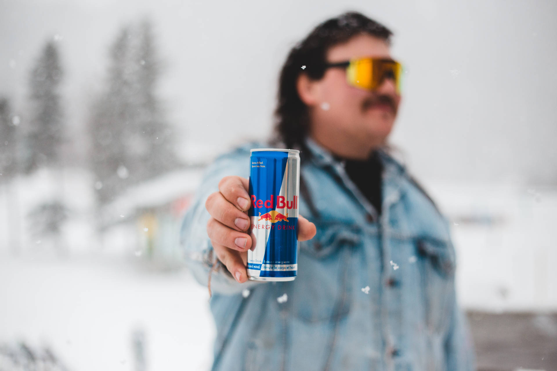 雪中男人手持紅牛罐，戴黃色護目鏡和牛仔外套。