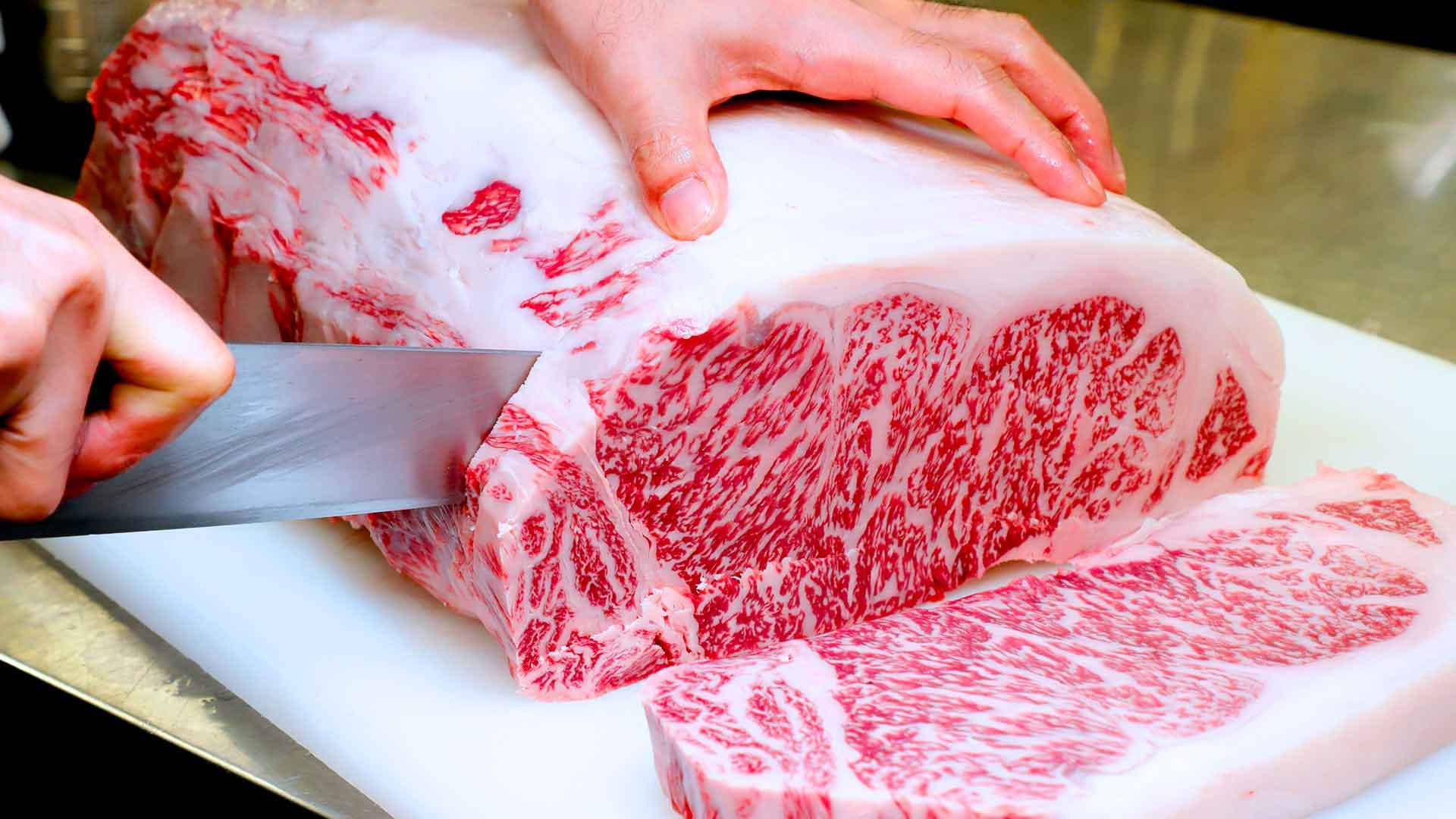 手在白色砧板上用刀切一大塊大理石紋烤肉。
