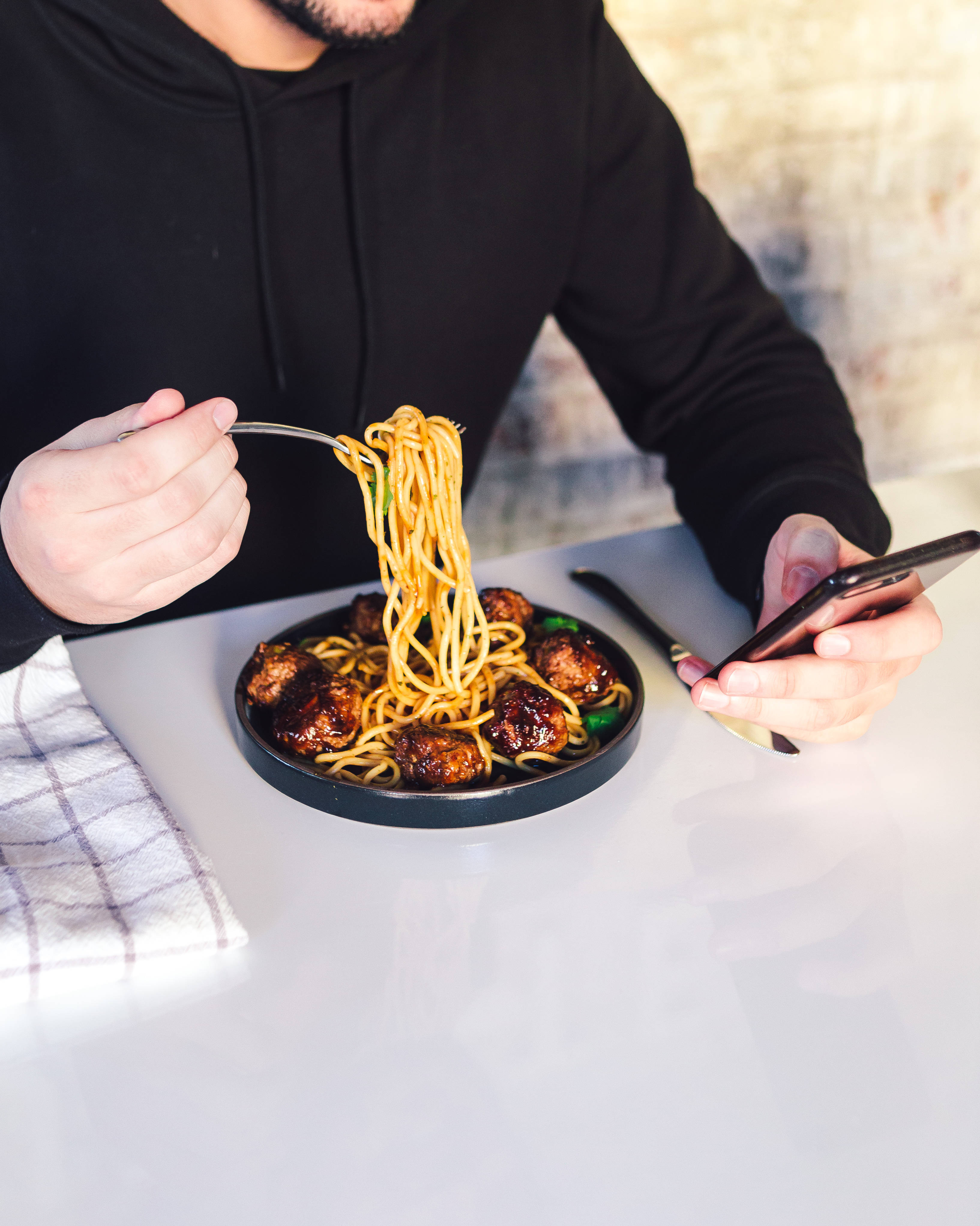 男子吃義大利麵 iPhone 壁紙 — 一個穿著黑色連帽衫的人，在白色桌子上從黑色盤子裡夾起帶有肉丸的麵條，同時滑動手機。