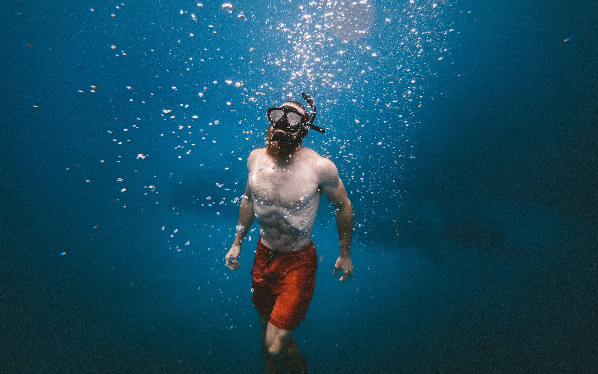 一位留鬍鬚的男子在藍色海水中浮潛，氣泡向水面上升。