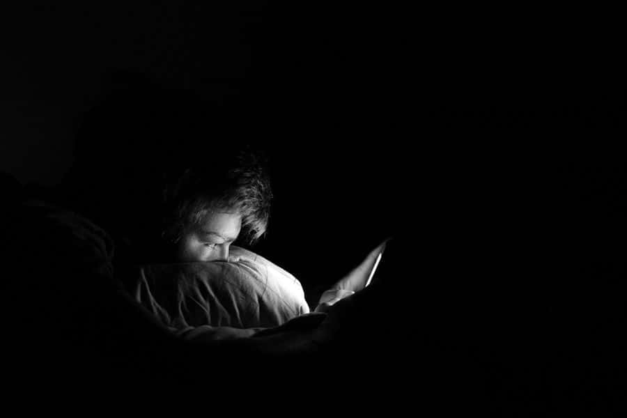 男子在黑暗房間前哭泣 壁紙 — 一個人蜷縮在床上被窩中，被黑暗房間中手持裝置的光芒照亮。