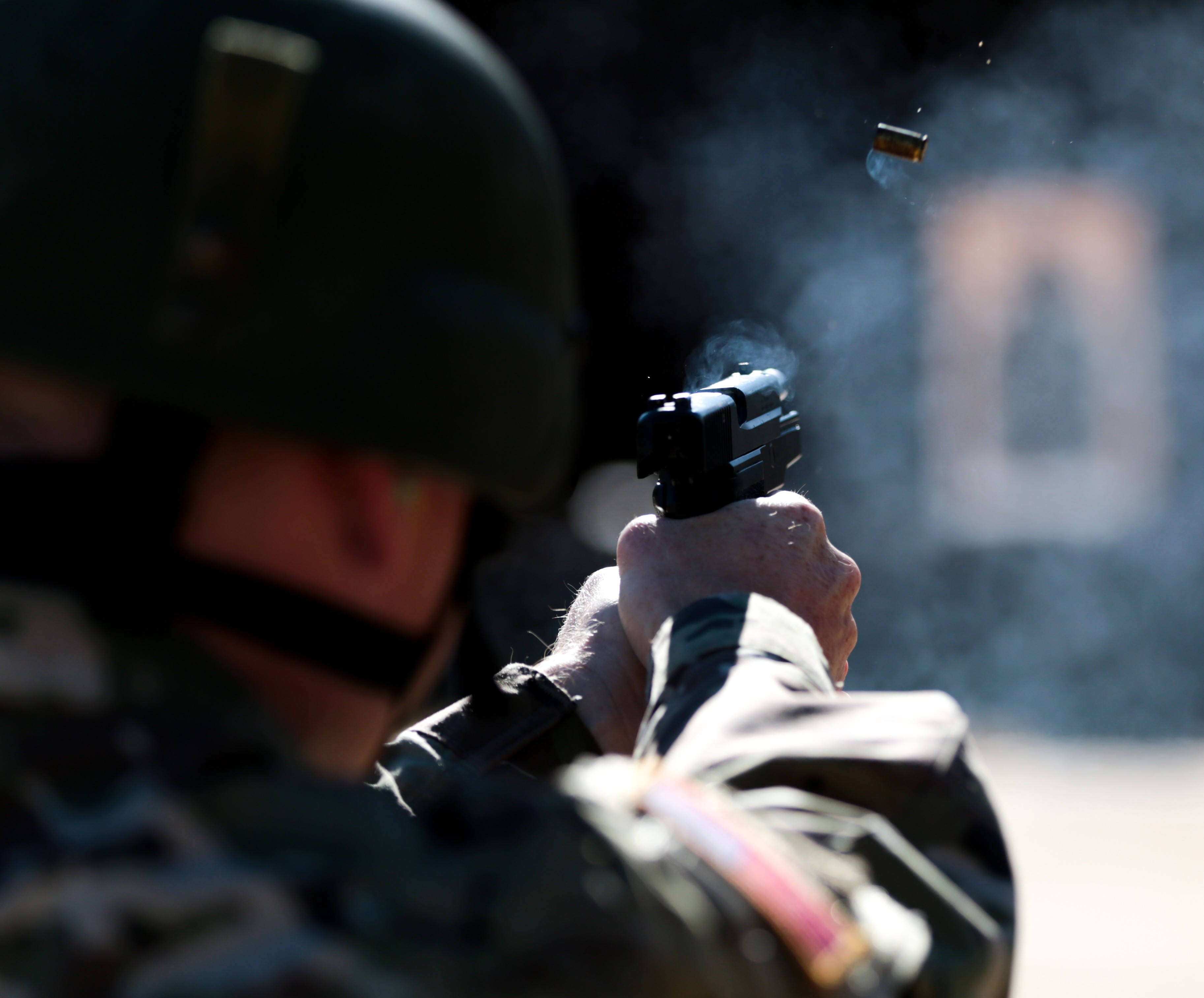 男子射擊特寫 壁紙 — 一名身穿迷彩服的士兵開槍射擊，一顆用過的彈殼在空中飛過，背景是模糊的目標。