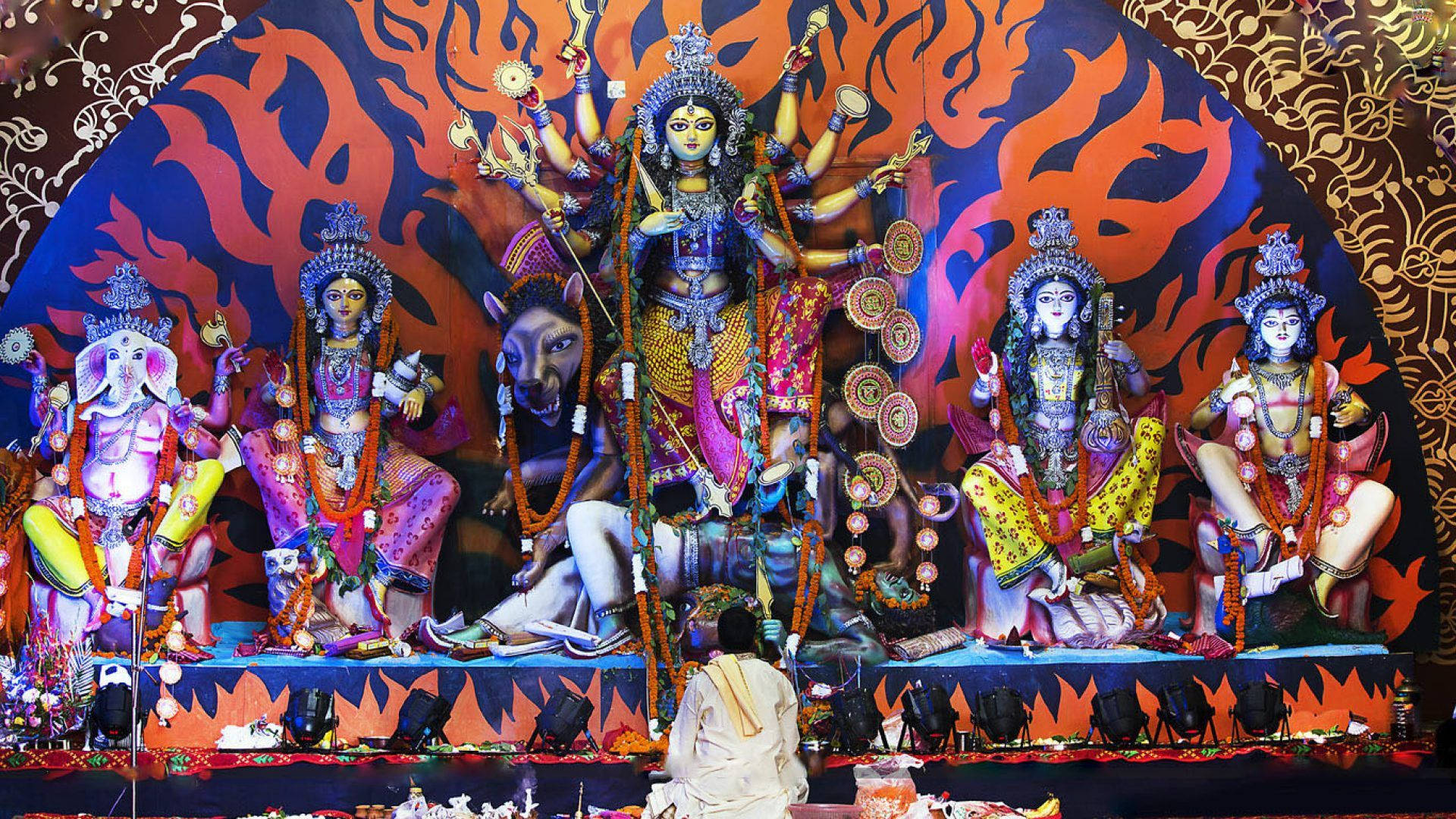 男子於 Durga Mata 高清 壁紙 — 一位信徒在熾熱的橙色和藍色背景下，向宏偉多彩的杜爾迦神像鞠躬，神像周圍環繞著她的神聖侍從。