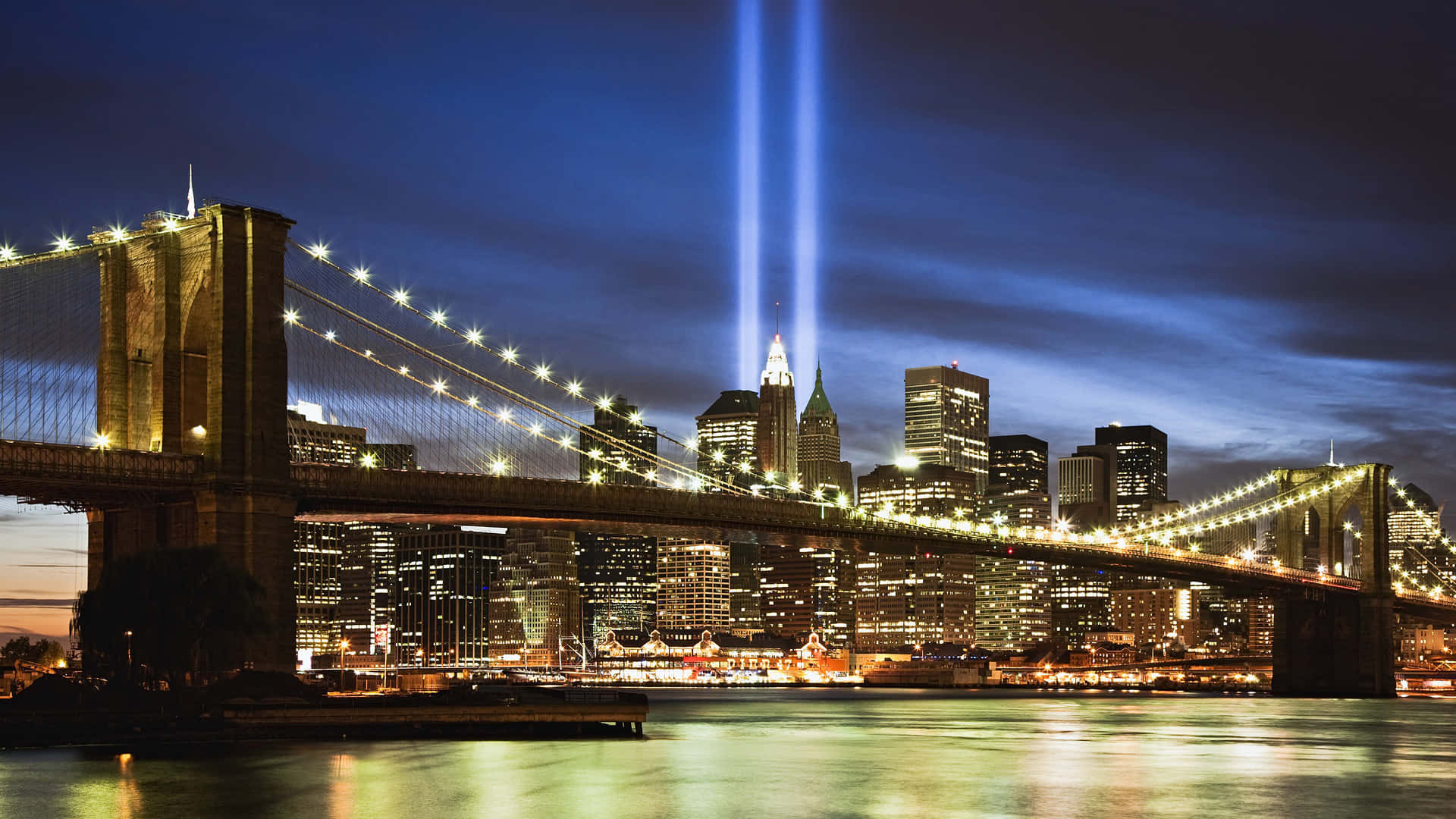 男子漫步紐約繁忙街頭 壁紙 — 夜晚的布魯克林大橋，Tribute in Light 光束刺穿曼哈頓天際線上的天空，倒映在水中。