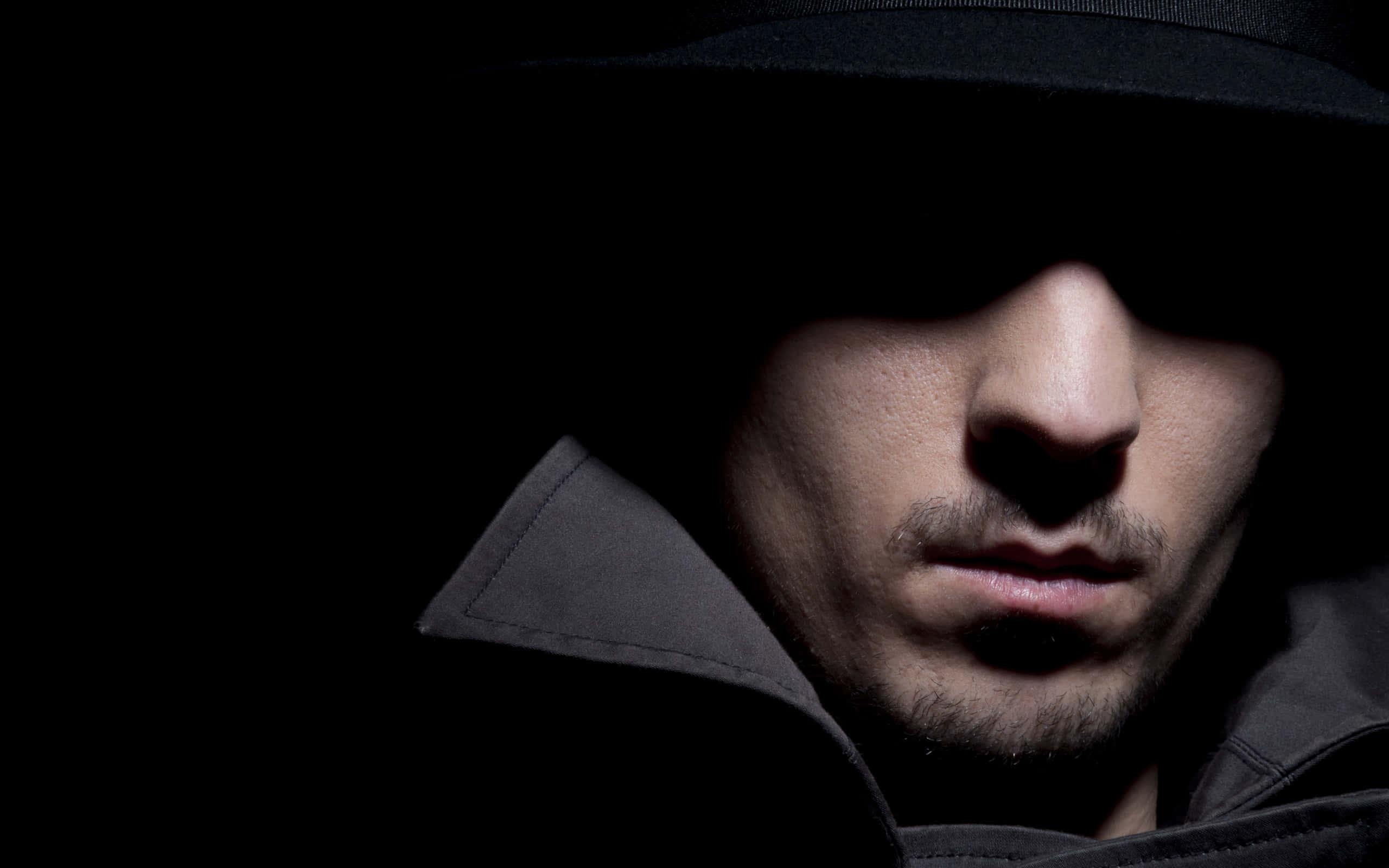 男子臉部隱於陰影中 壁紙 — 一名戴黑色軟呢帽、穿 trench coat 的男子，在戲劇性的明暗對比燈光下從黑暗中凝視。