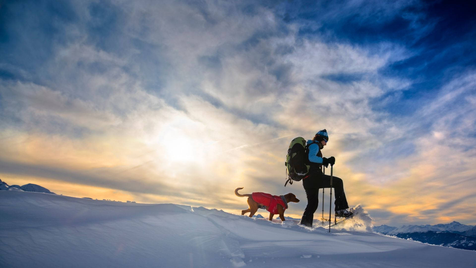 男子與愛犬雪鞋健行 壁紙 — 一位穿著紅色外套的遠足者帶著狗在日落時分，在戲劇性的雲彩和金色光芒中攀登白雪覆蓋的山脊。