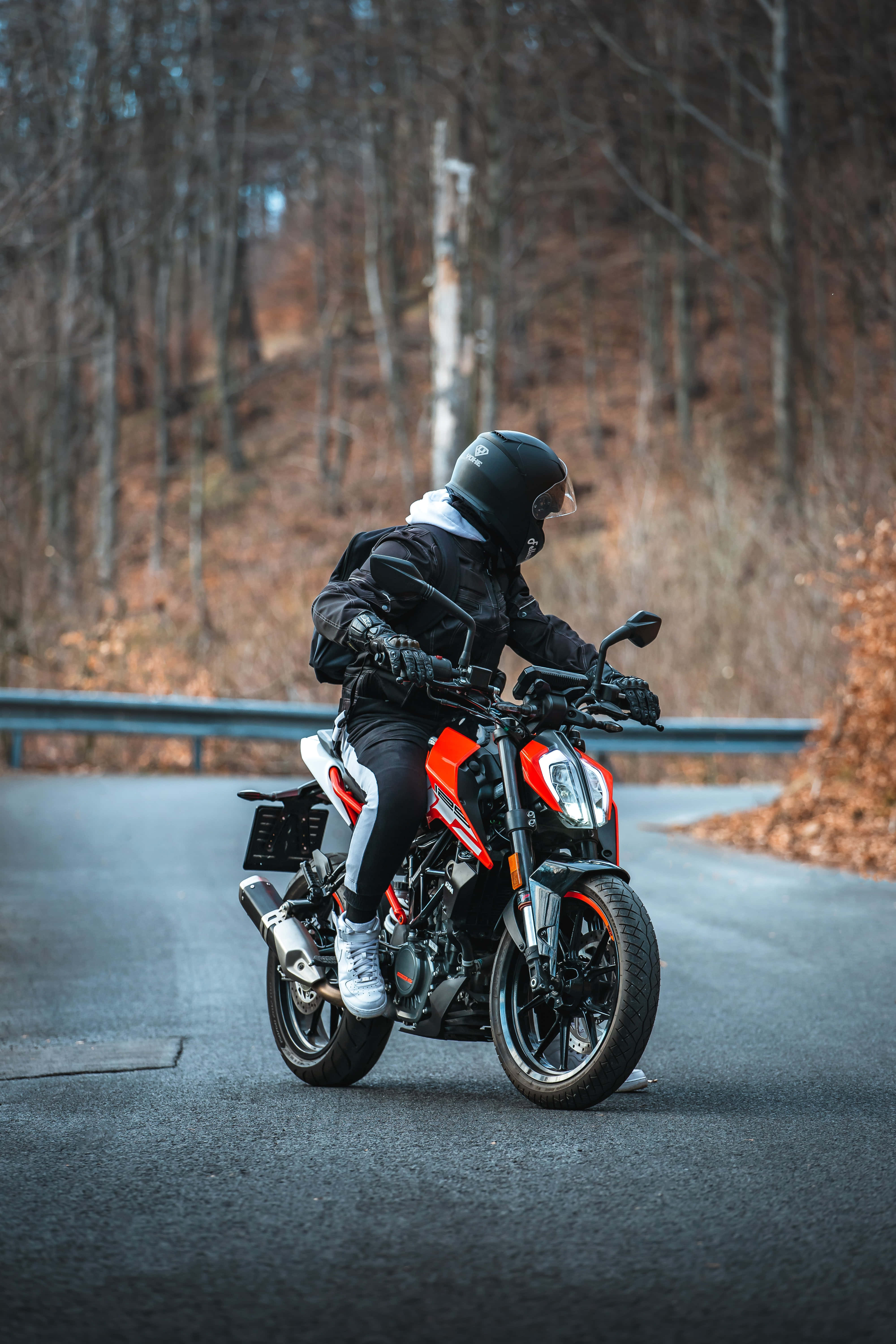 男子騎乘 KTM Duke 390 機車照片 壁紙 — 一名全副裝備的電單車手在兩旁光禿樹木的蜿蜒森林道路上騎著一輛紅黑配色的跑車。