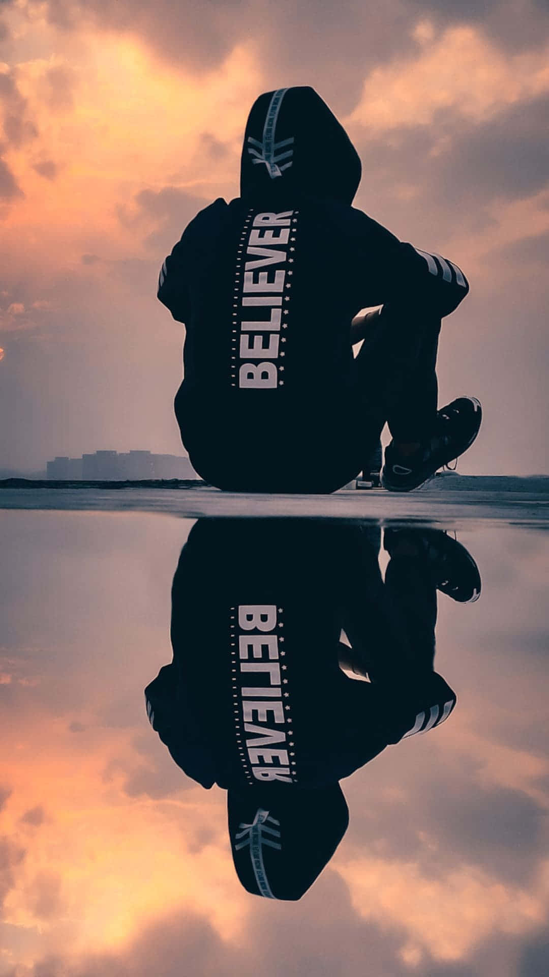 穿著 believer 連帽衫的人坐在水邊，帶有日落倒影的剪影。