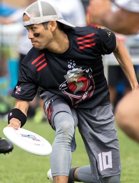 男運動員右看持飛盤Android Ultimate Frisbee背景 壁紙 — 一名專注的男性終極飛盤選手，身穿黑色和紅色裝備，在草地上向前衝刺，手持白色飛盤。